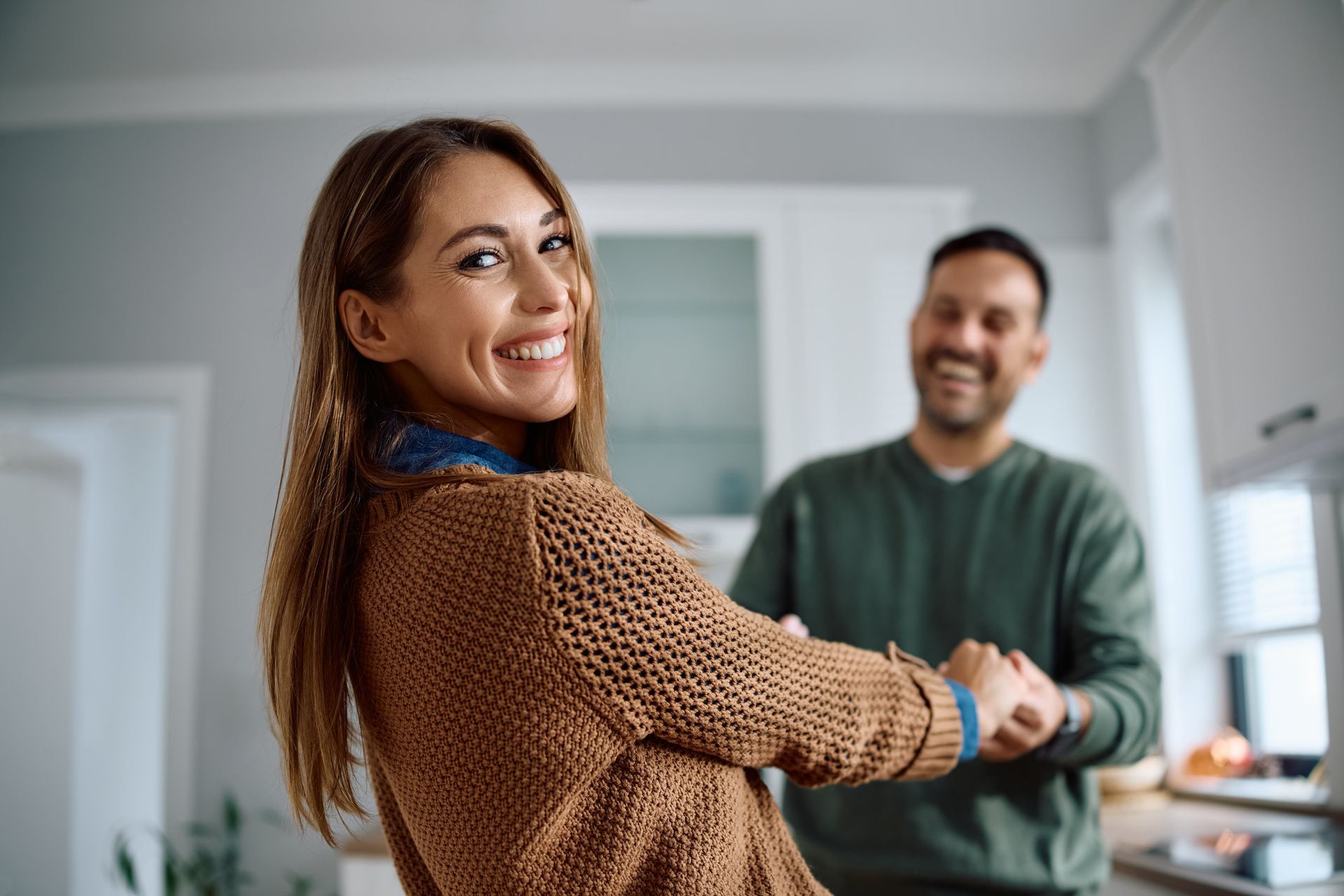 Woman in brown sweater smiles and holds hands with smiling person in green sweater in a kitchen.