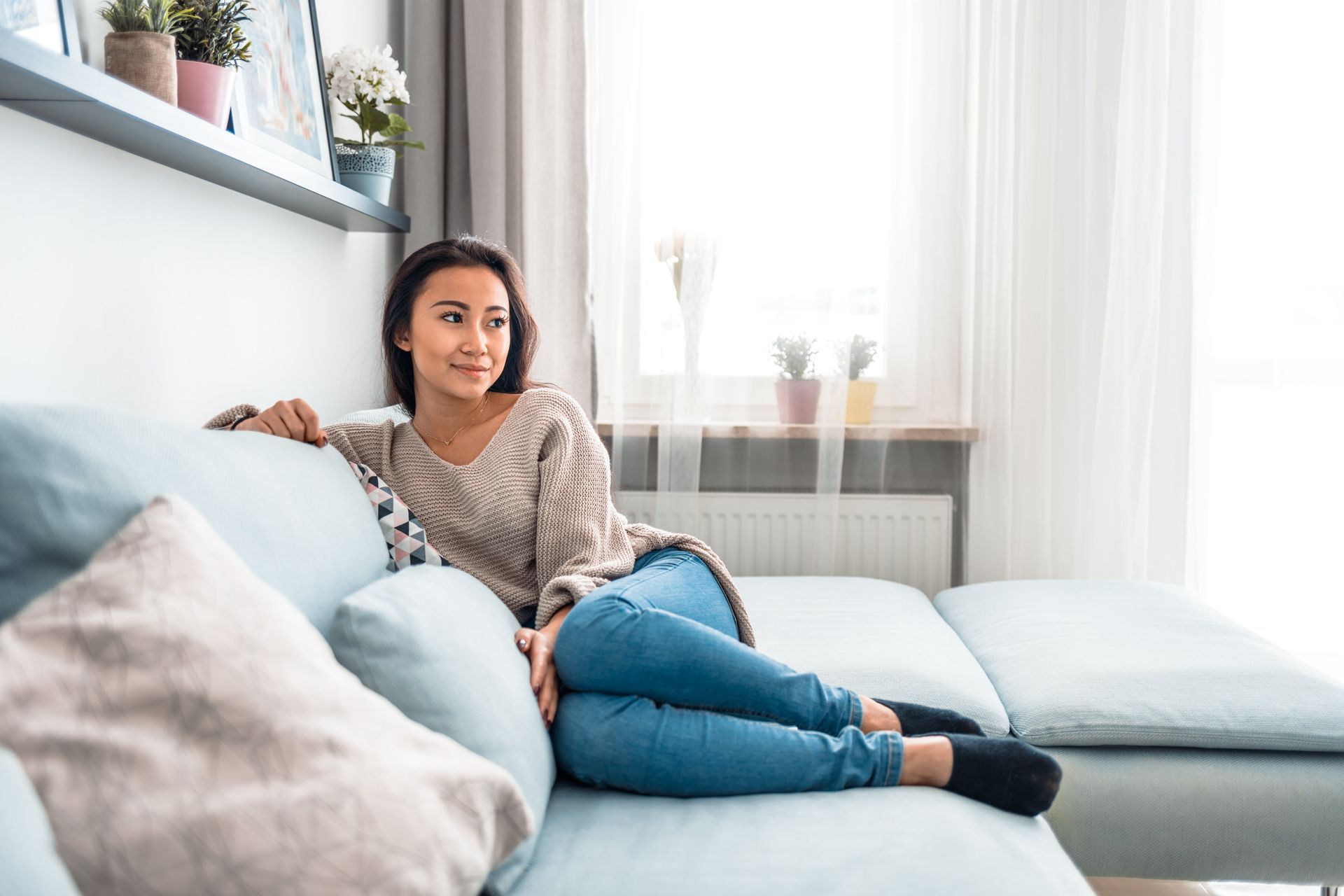 Woman relaxing on a blue couch, looking out window. Wearing a sweater and jeans, socks on feet, in a bright room.