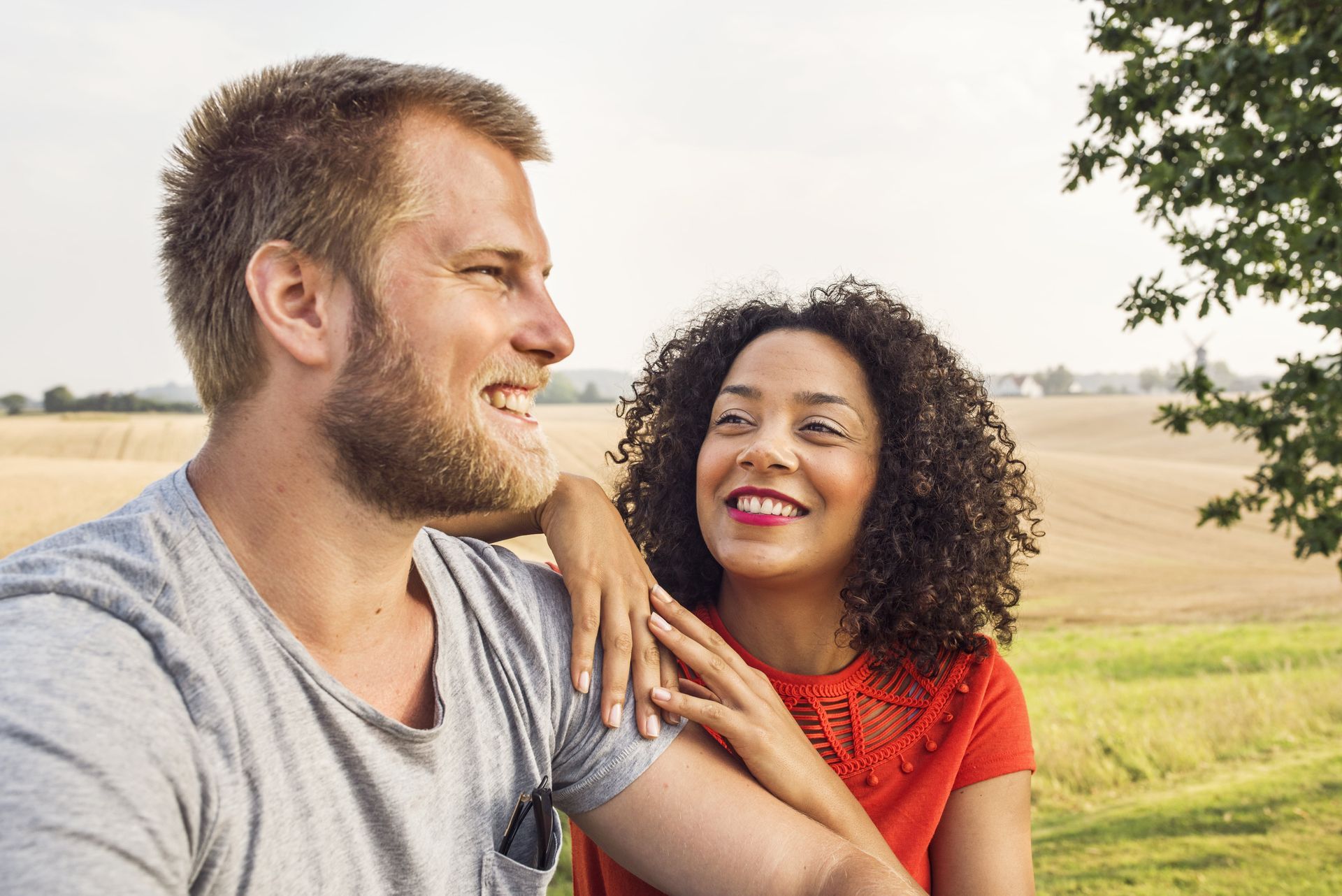 Man smiles, woman's hand on his shoulder. They look at each other, outdoors near a field.