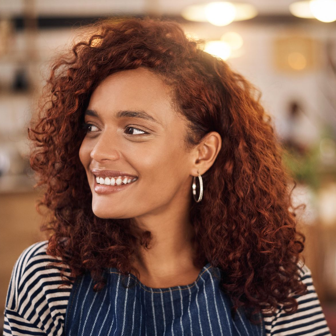 Woman with curly reddish hair smiling, wearing a striped shirt and apron, looking to the side in a cafe.