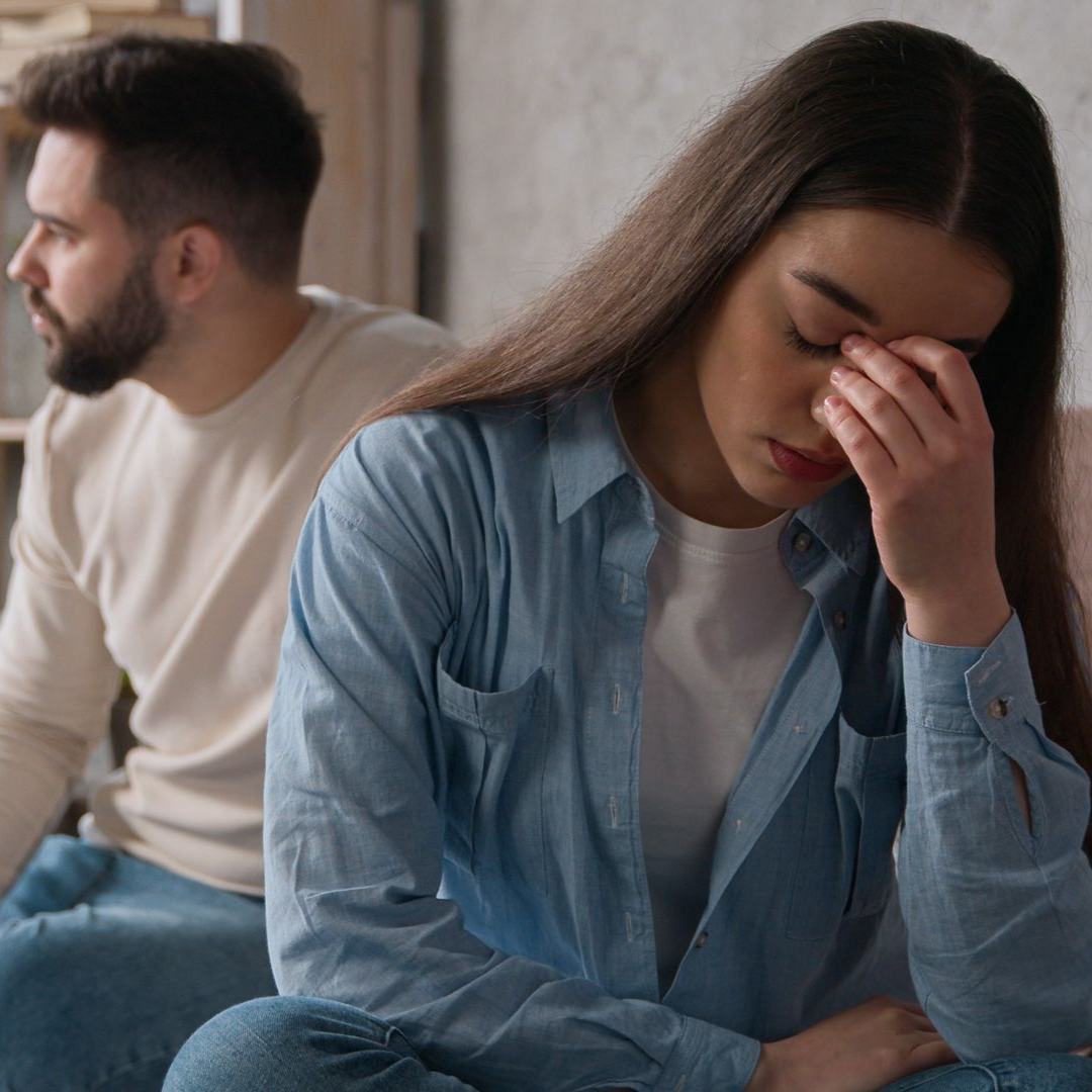 Woman with hand on forehead, looks distraught while man looks away; seated indoors.