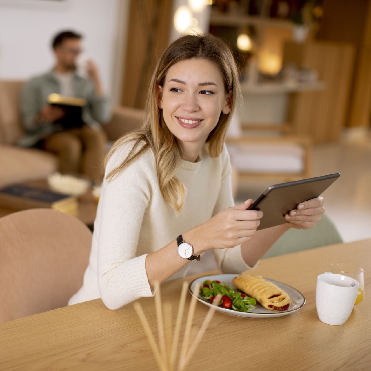 Woman smiling, holding tablet at table with food; man reading in the background.