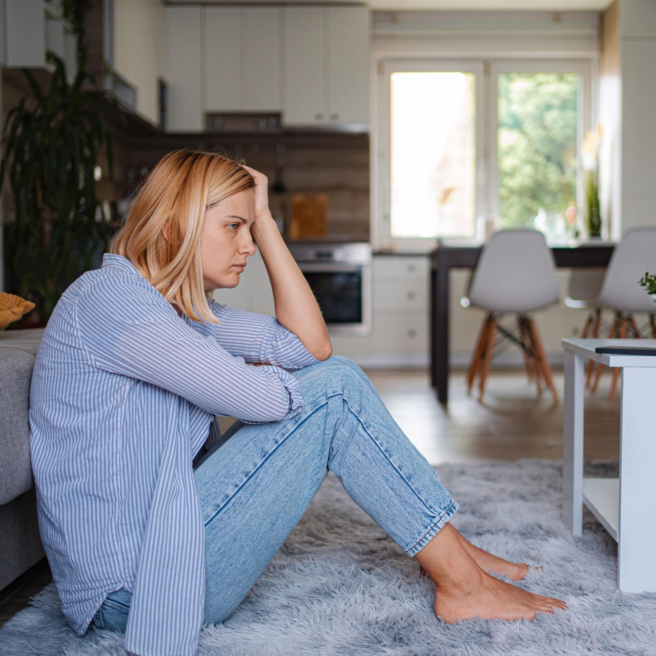 Woman with blonde hair sits on a rug, head in hand, looking sad. Living room with open kitchen in background.