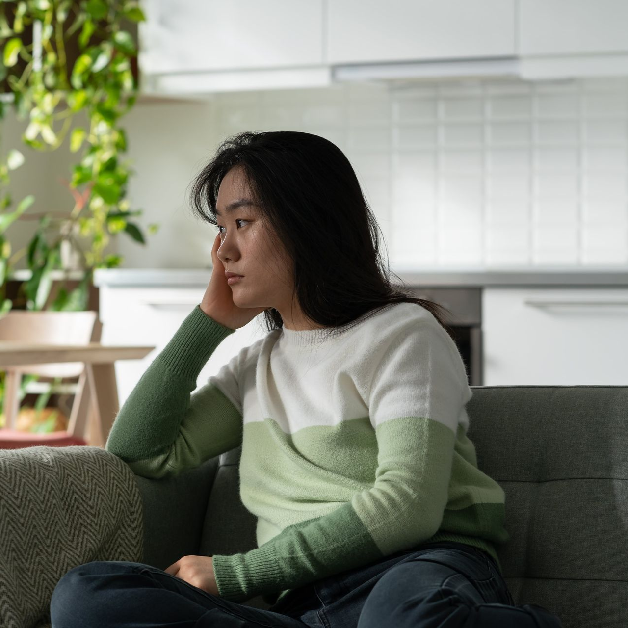 Woman sitting on a couch, looking pensive in a living room with plants and a bright kitchen in the background.