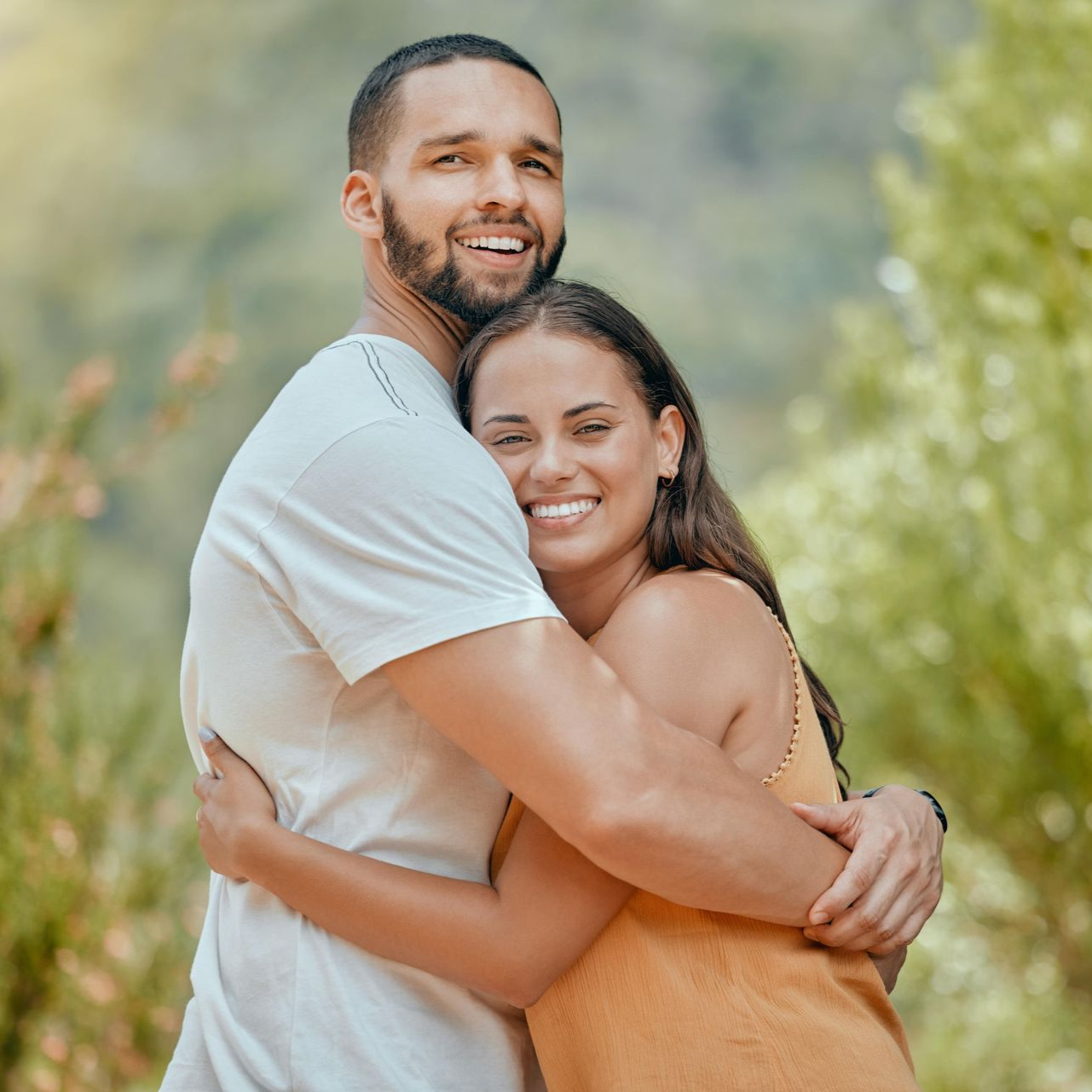 Couple hugging and smiling outdoors. The man wears a white shirt, the woman an orange dress.