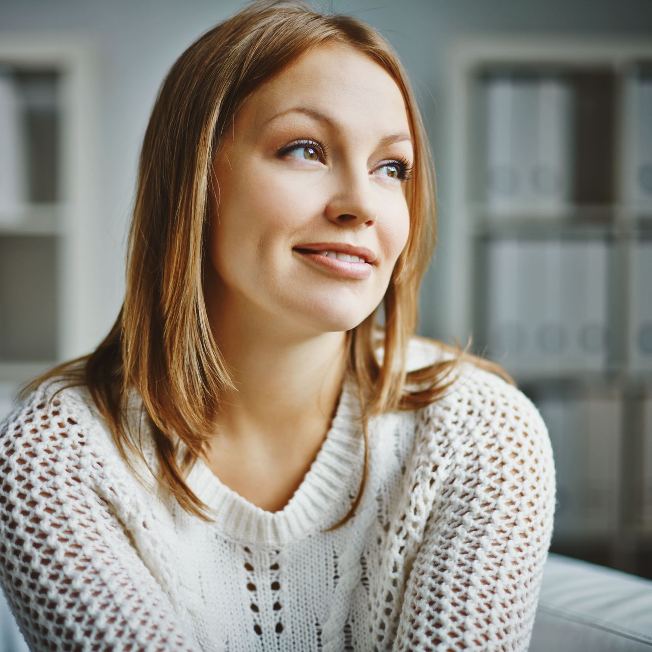 Woman in white sweater looks off to the side, smiling. Inside setting with blurred background.