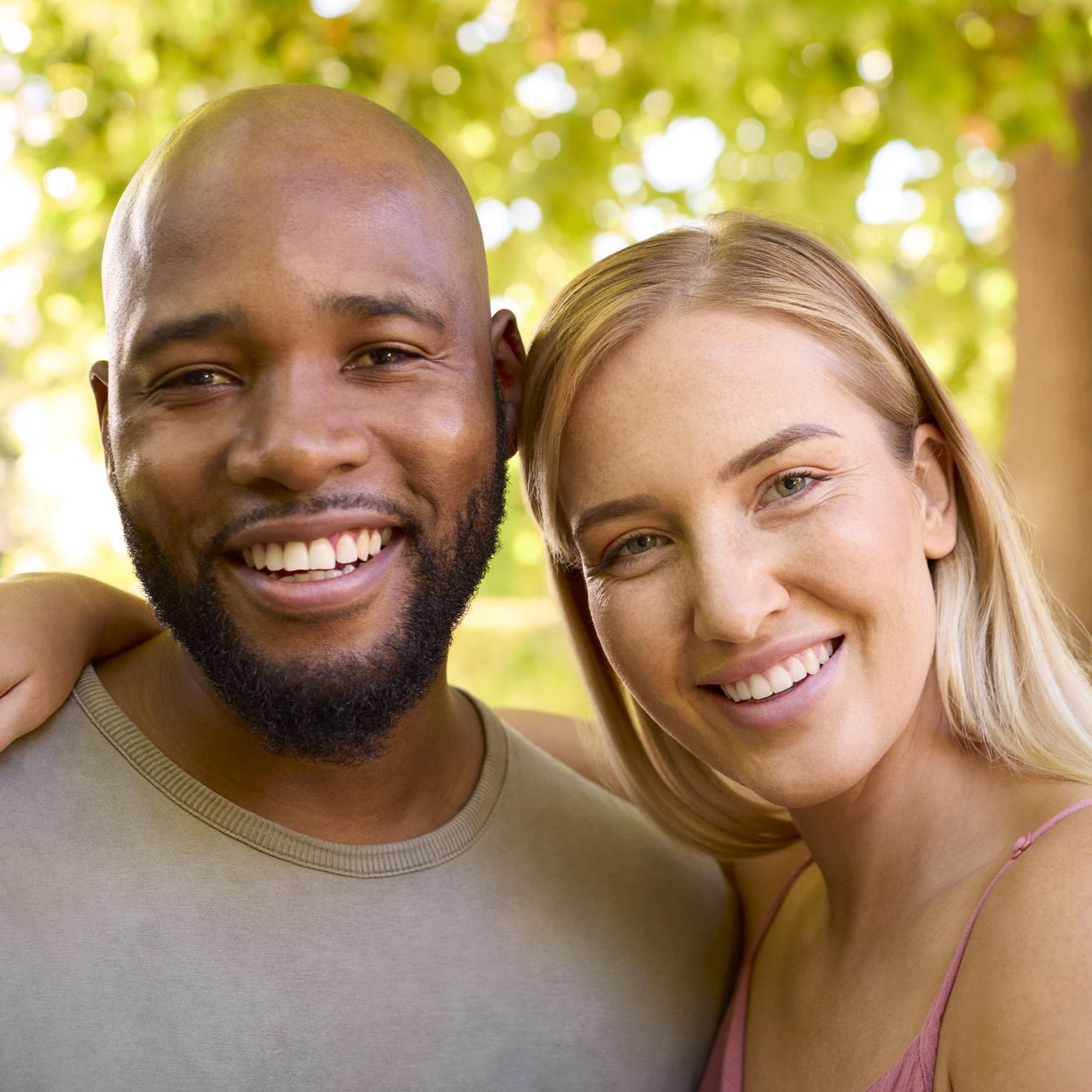 Man with beard and blonde woman smile, embracing outdoors.