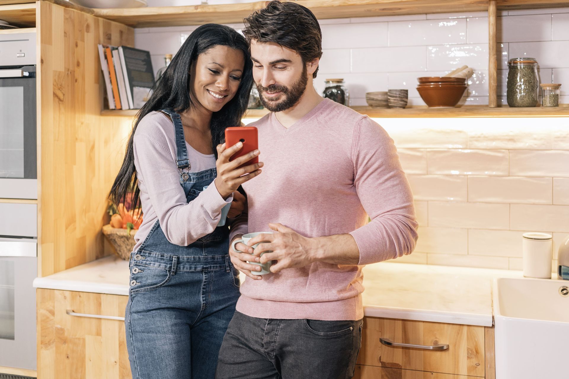 A couple looking at a phone together in a kitchen. They are smiling, with light wood cabinets.