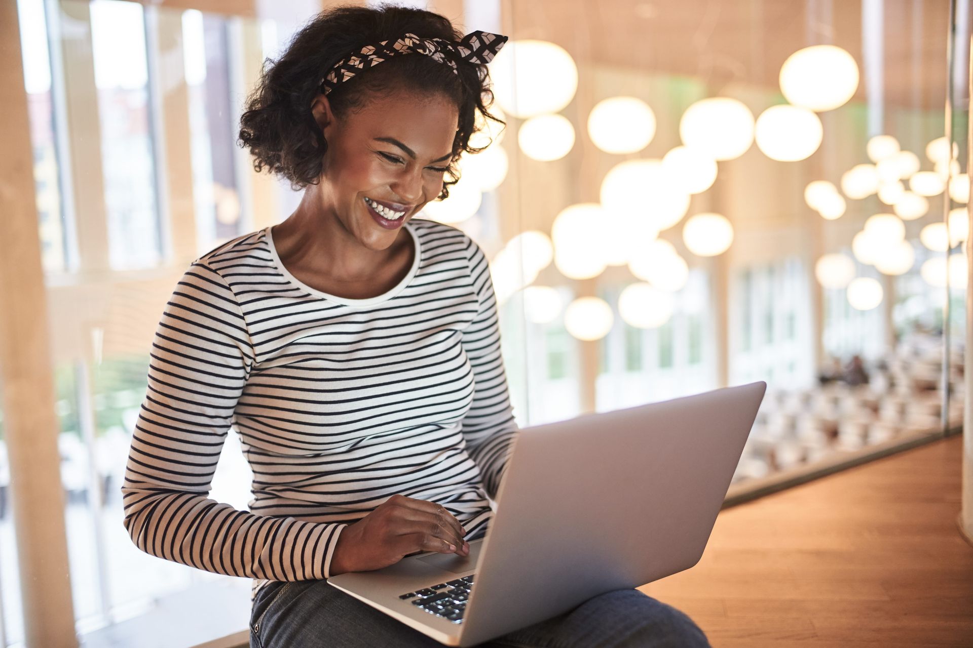 Woman smiling, using a laptop. Wearing striped shirt, headband. Bright office with lights in background.
