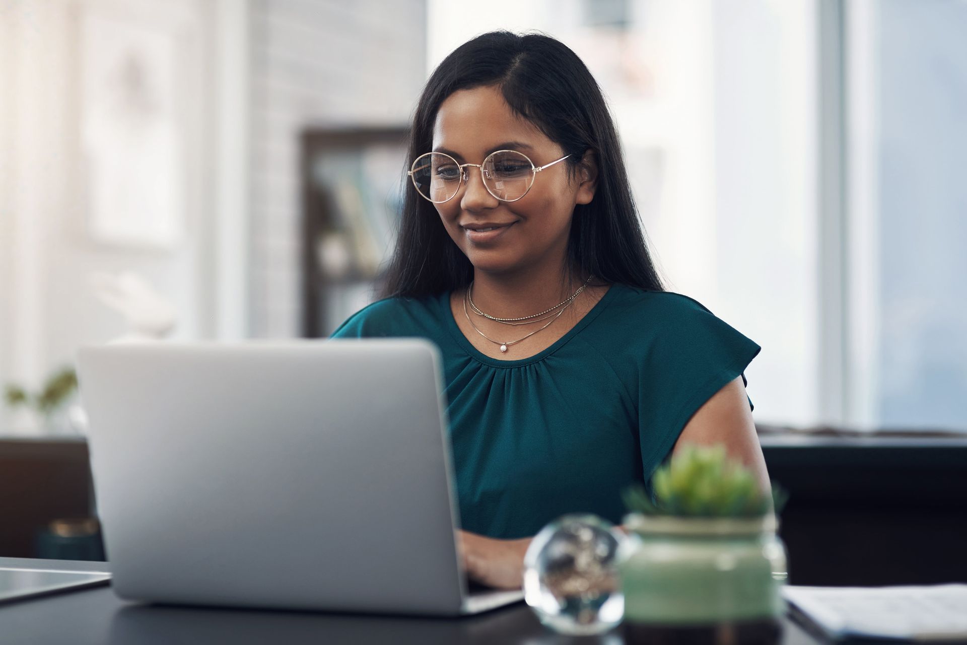 Woman with glasses working on a laptop, smiling at her desk.