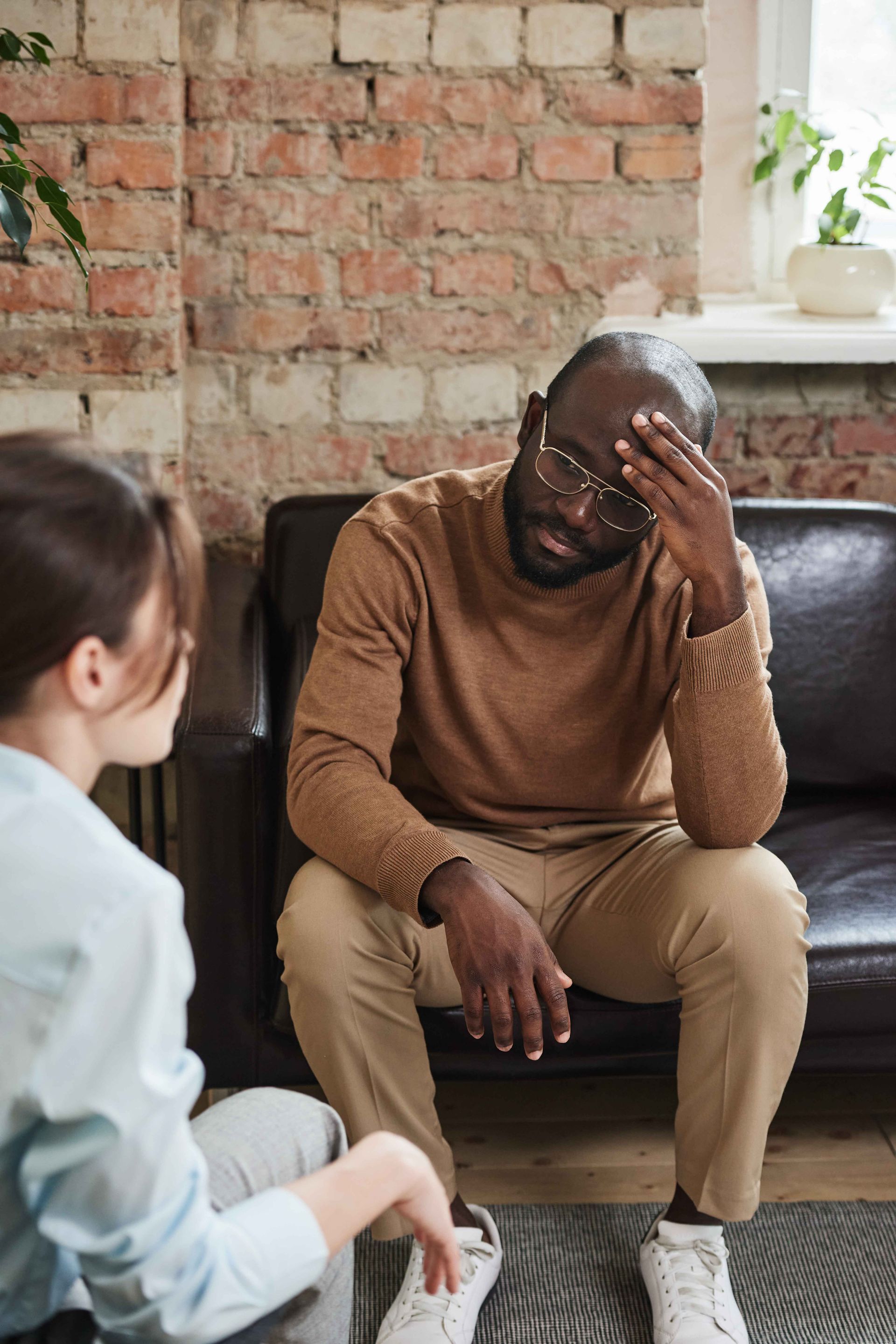 Man seated on a couch with hand on forehead, talking to a person, indoors.