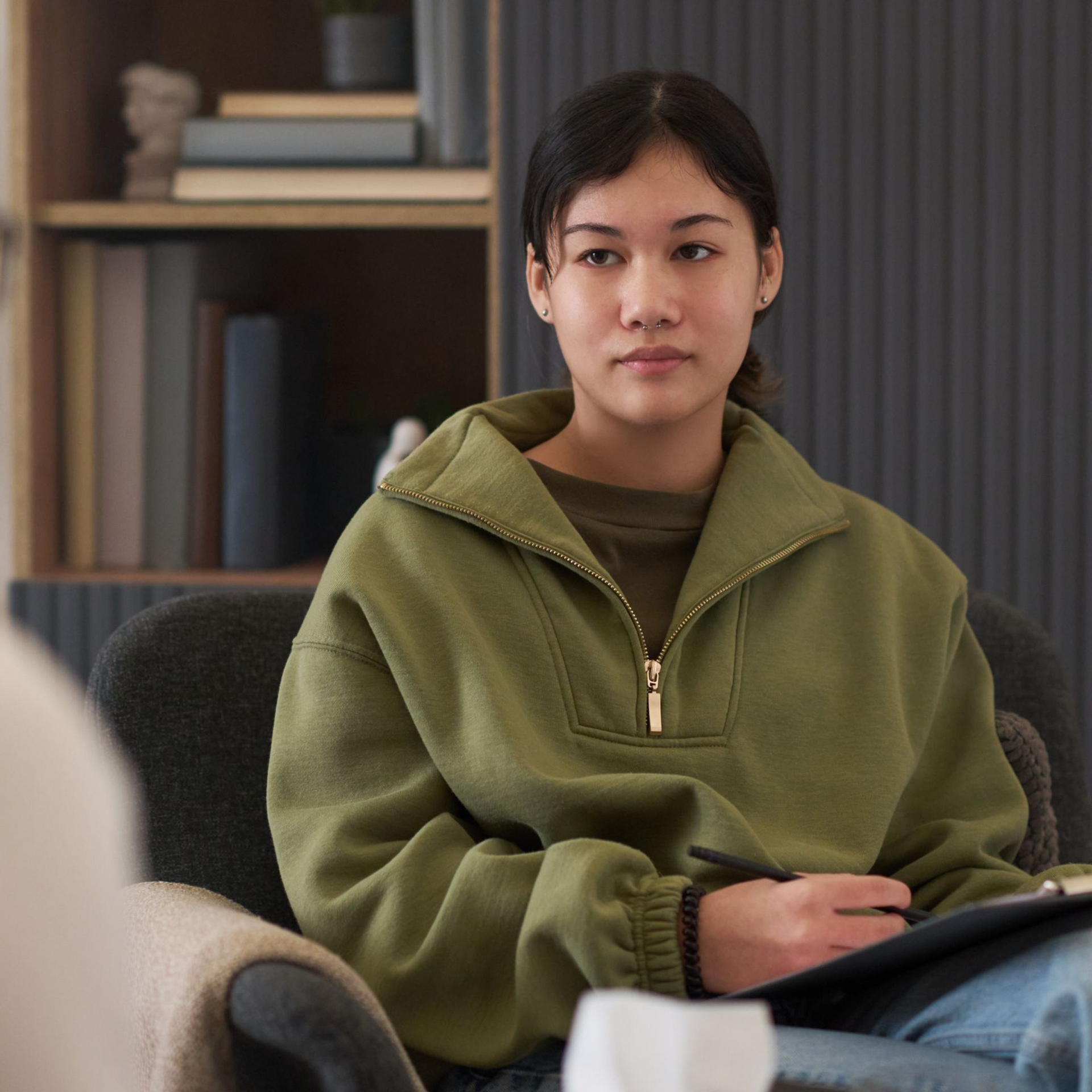 Woman seated, looking forward, in a therapy session. Plant, window, and person in blurred foreground.