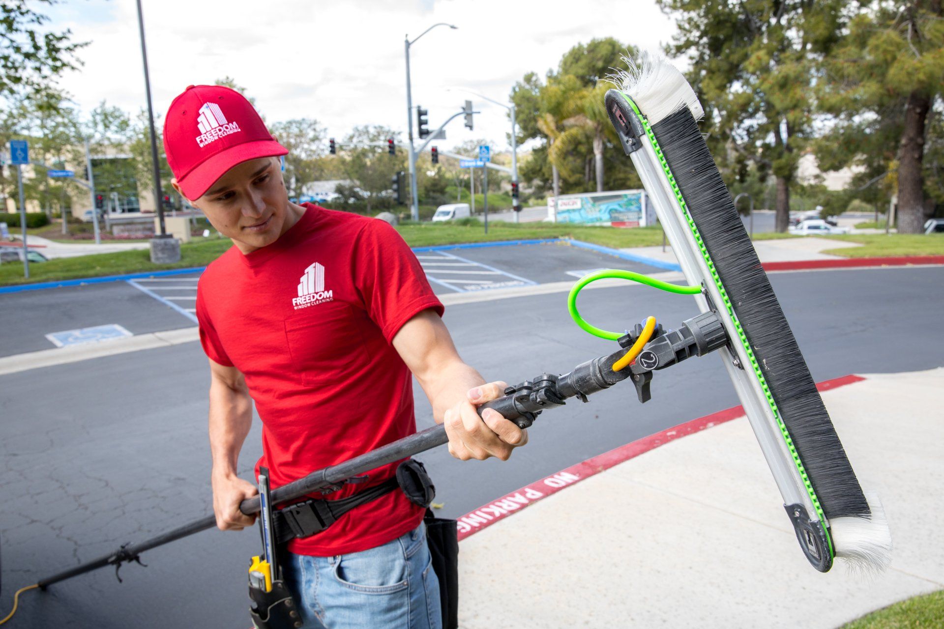 Man in red shirt and cap using a window cleaning tool on the sidewalk near a street.
