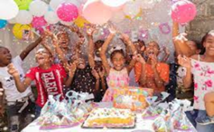 A group of children are standing around a table with balloons and confetti.