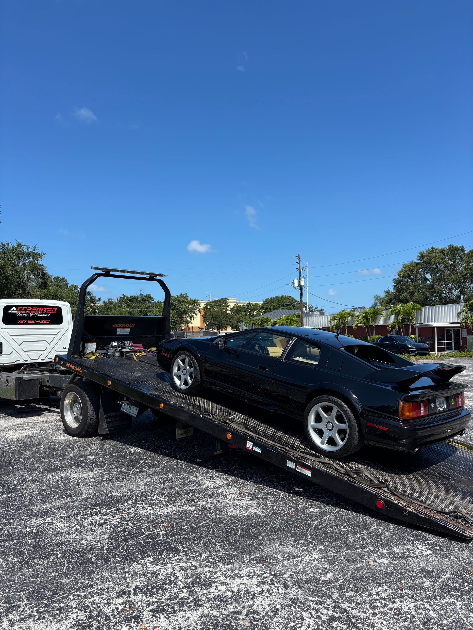 Black Lotus sports car being towed on a flatbed tow truck on a sunny day.