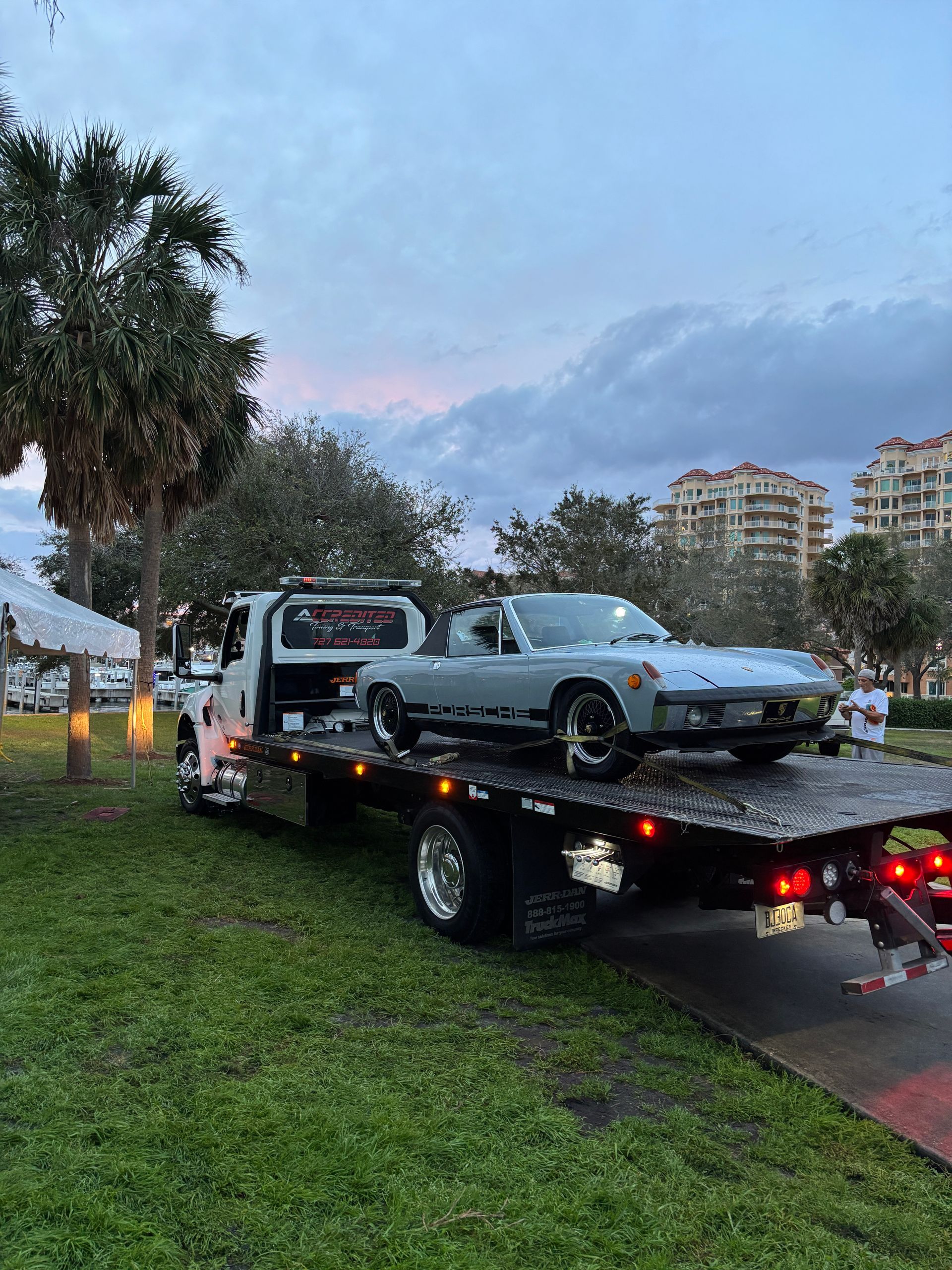 Porsche 914 convertible loaded on a tow truck, parked on grass near a building and palm trees, under cloudy skies.