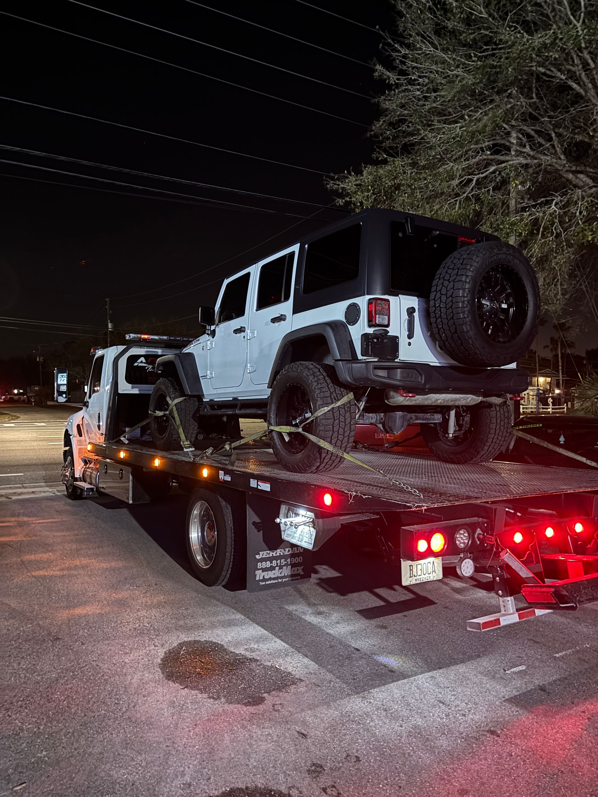 A light gray Jeep Wrangler loaded on a flatbed tow truck at night.