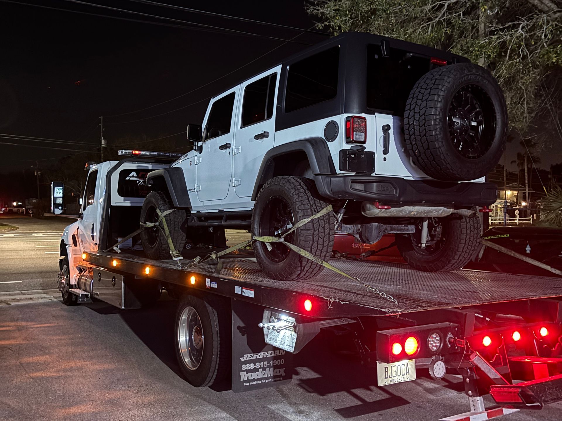 White Jeep SUV on a tow truck at night, secured with straps, with red lights visible.