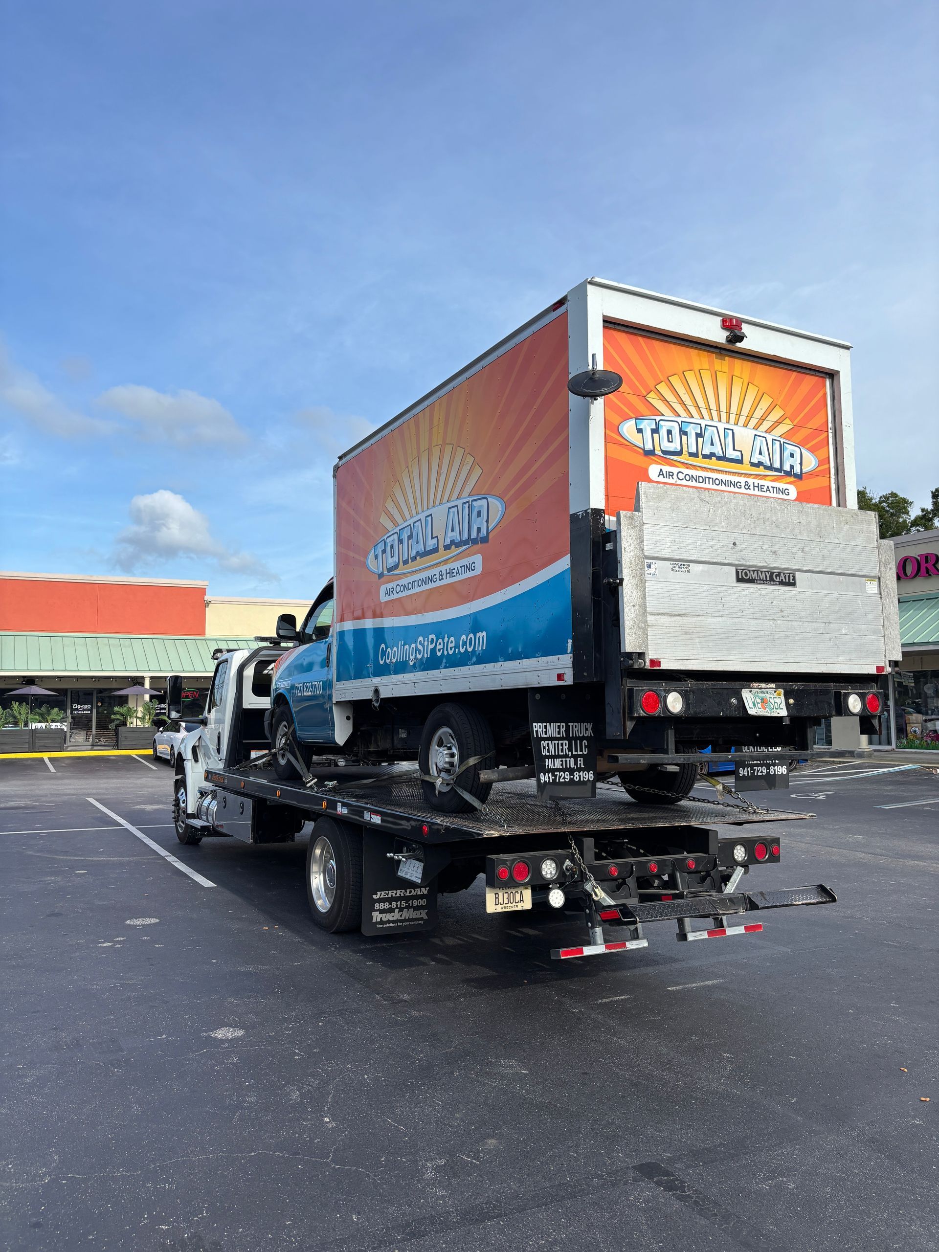 A tow truck carrying a light blue truck with a colorful advertising box in a parking lot under a cloudy sky.