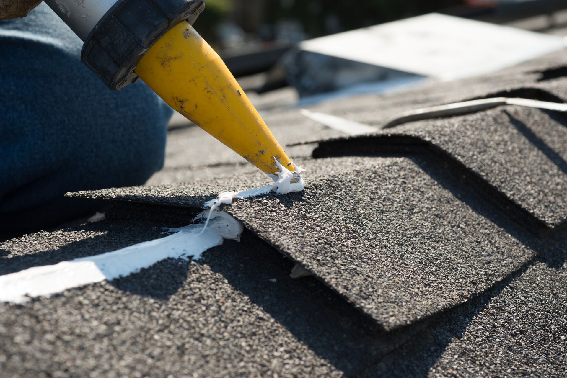 A person uses a caulk gun to apply white sealant along a seam between asphalt roof shingles. A person uses a caulk gun to apply white sealant along a seam between asphalt roof shingles.