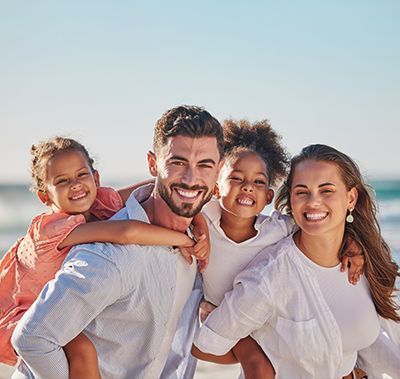 Family of four smiling on a beach, two children piggybacking on adults.