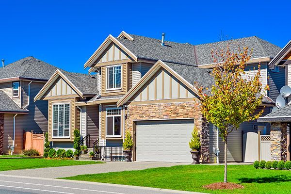 Two-story house with gray roof, tan trim, stone accents, and a green lawn under a blue sky.