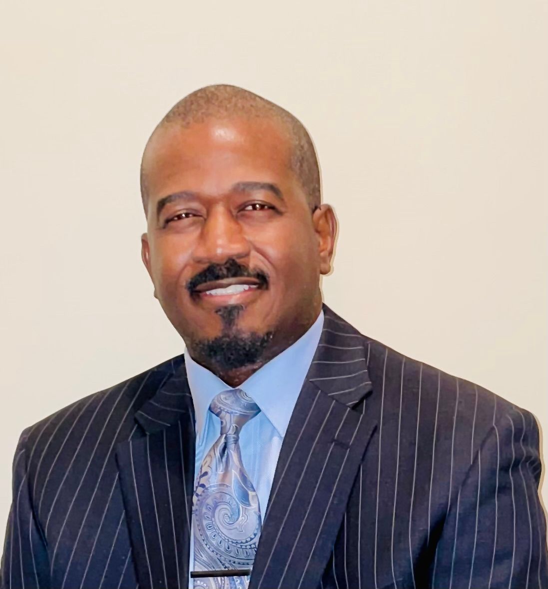 Man in a pinstripe suit and tie smiles, chest up portrait, beige background.