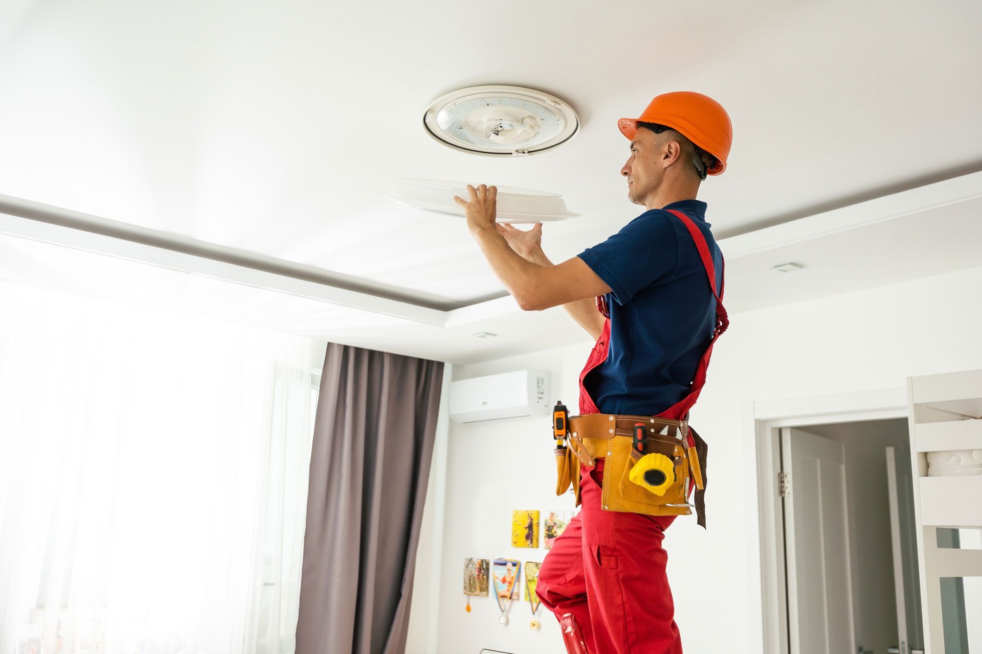Man in work clothes on a ladder, installing a light fixture on a white ceiling.