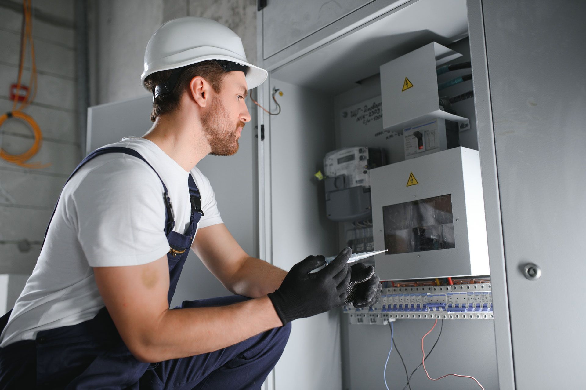 Electrician in a white hard hat inspects wiring inside an electrical panel.