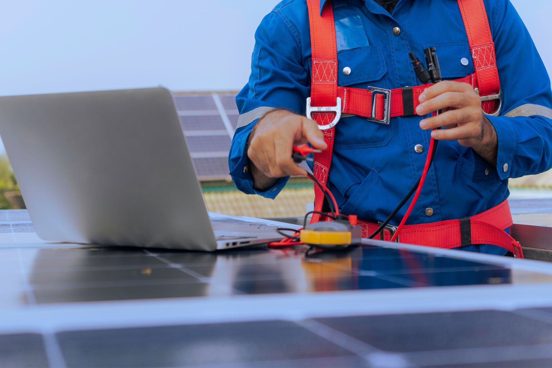 Solar panel technician using a multimeter on panels while wearing a safety harness and using a laptop.