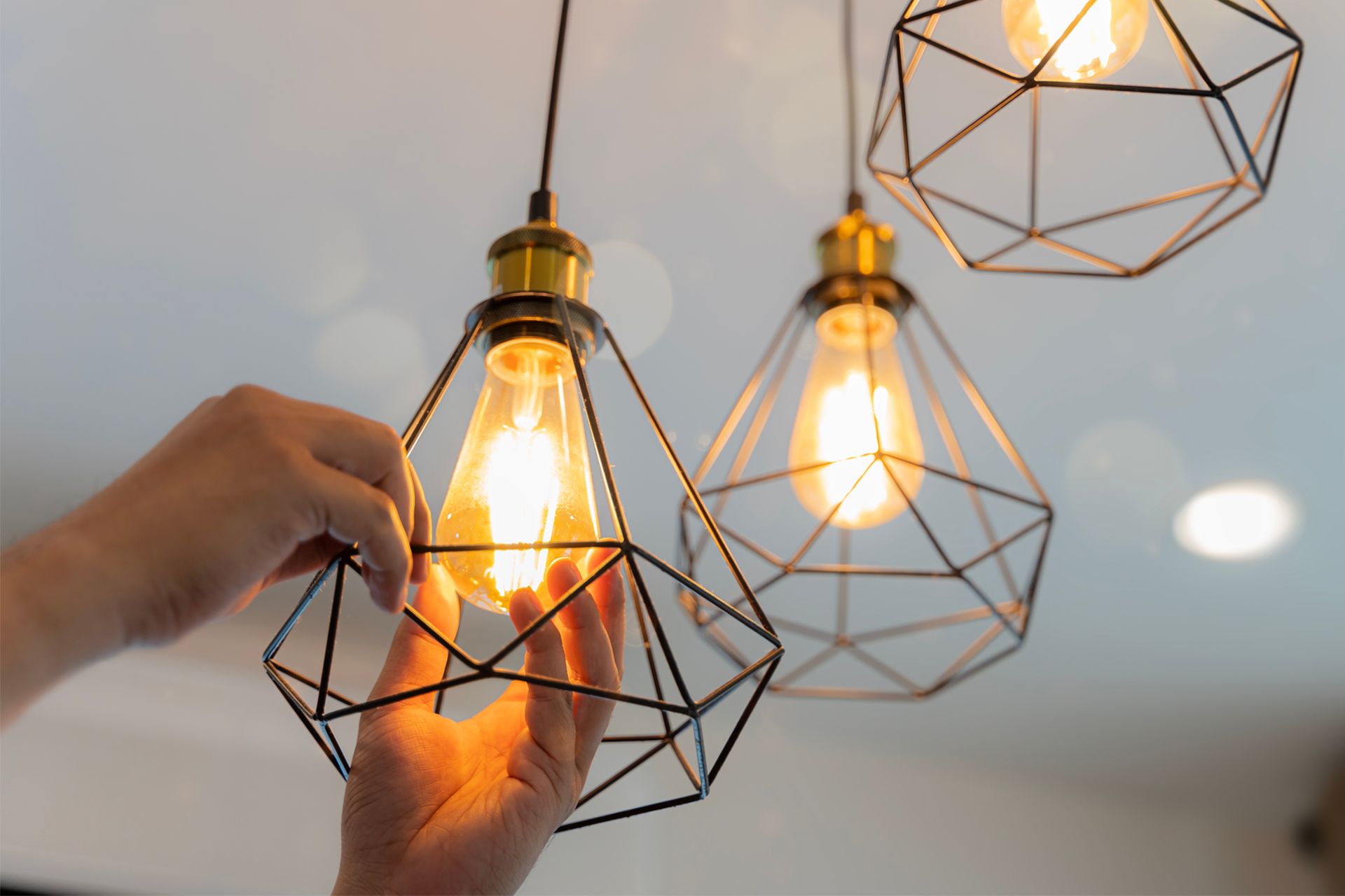 Person's hand adjusting a lit pendant light with a geometric wire frame.