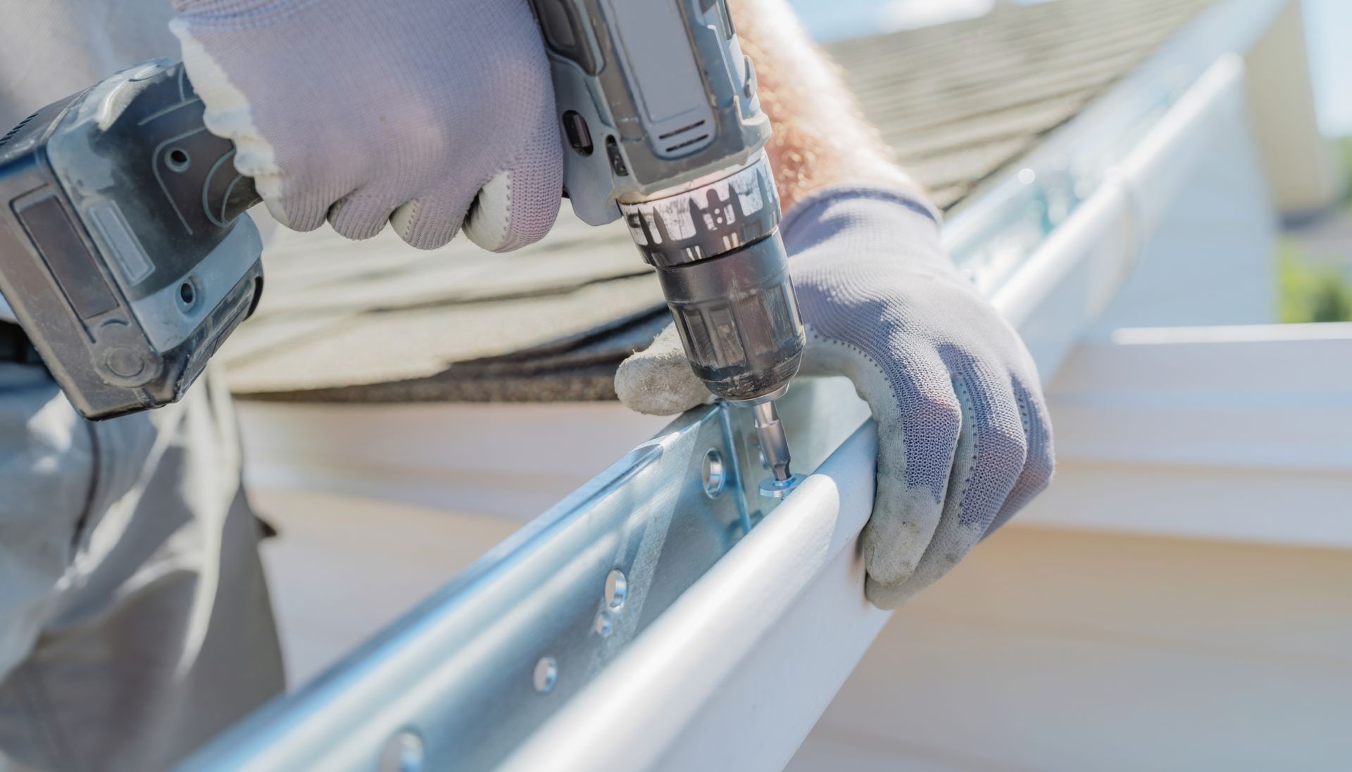 Person in gloves using a drill to install a white gutter on a house roof.