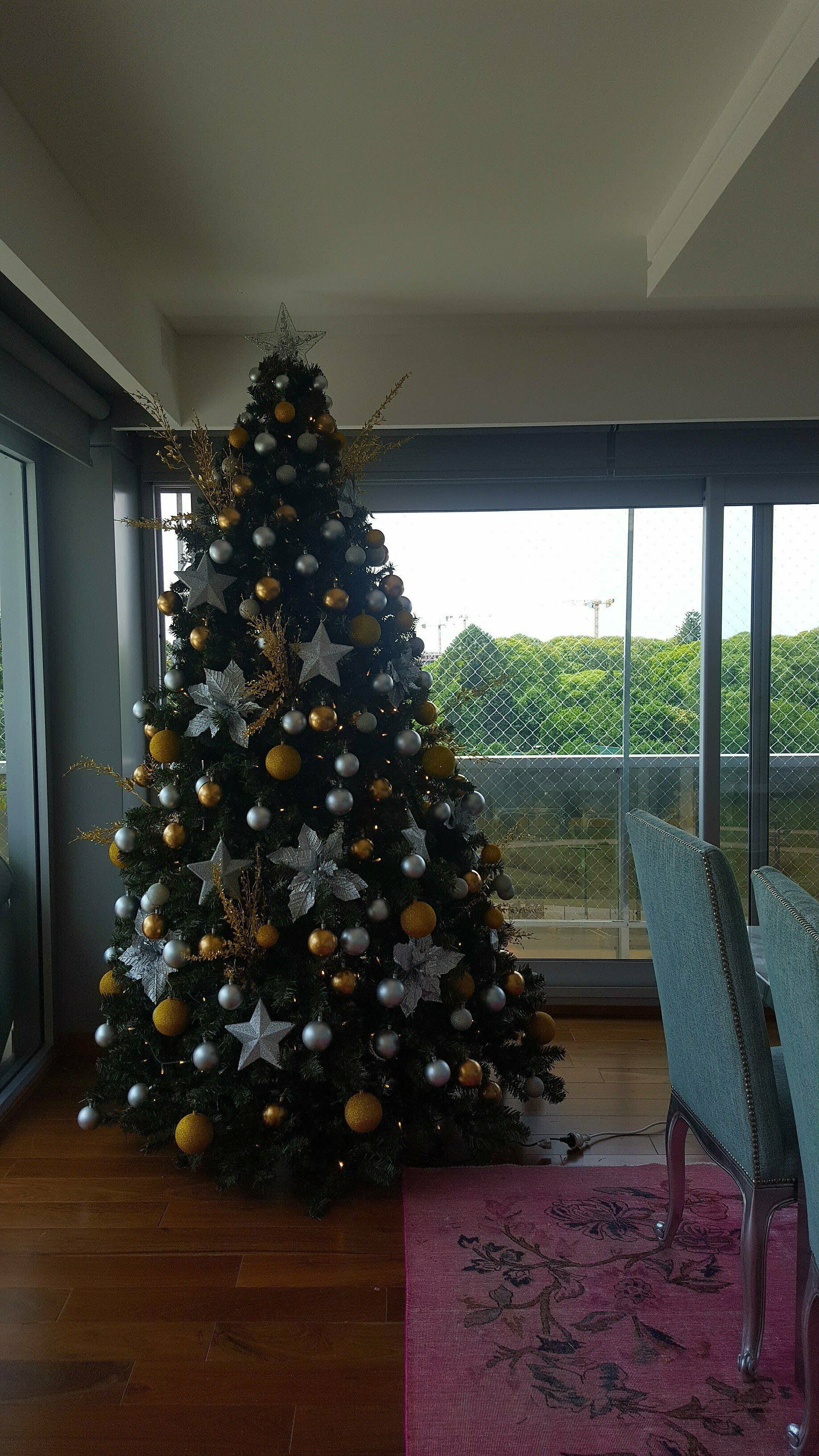 Árbol de Navidad decorado junto a una ventana en una sala de estar, con una alfombra rosa y sillas cerca.