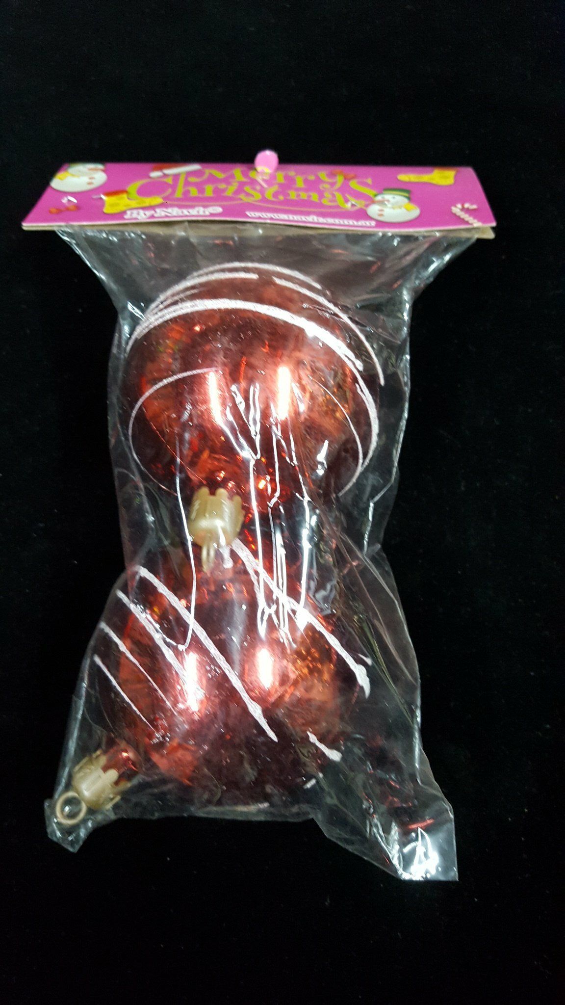 Muñeca o figurita decorativa roja en una bolsa de plástico transparente con una etiqueta rosa sobre fondo negro.