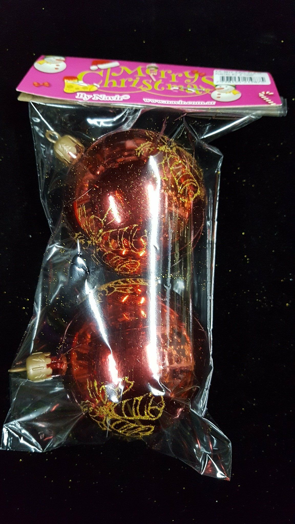 Un adorno navideño rojo y dorado envuelto en un embalaje de plástico transparente con una tarjeta de cabecera rosa.