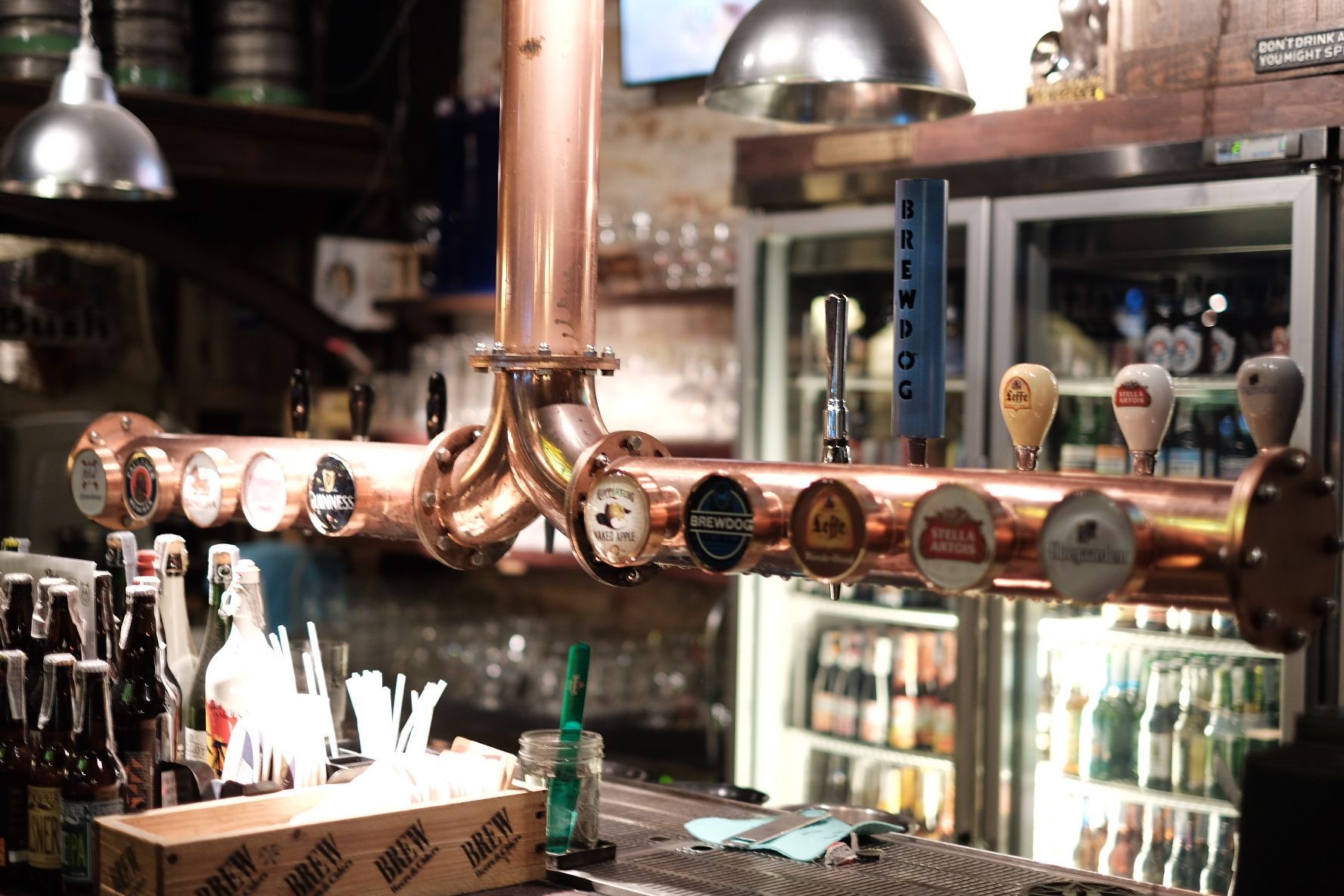Copper beer taps at a bar with various beer labels and a refrigerated display in the background.