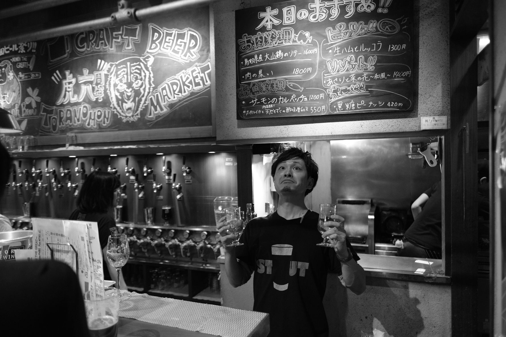 Man holding up two glasses in a bar, with beer taps and menu boards in the background.