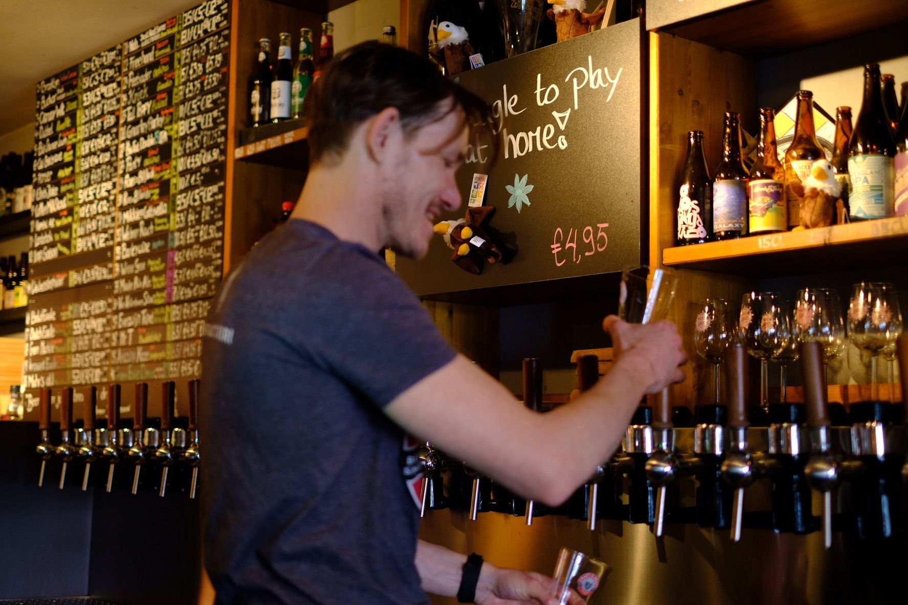 Bartender pouring beer from a tap, smiling, at a bar with beer bottles on shelves.