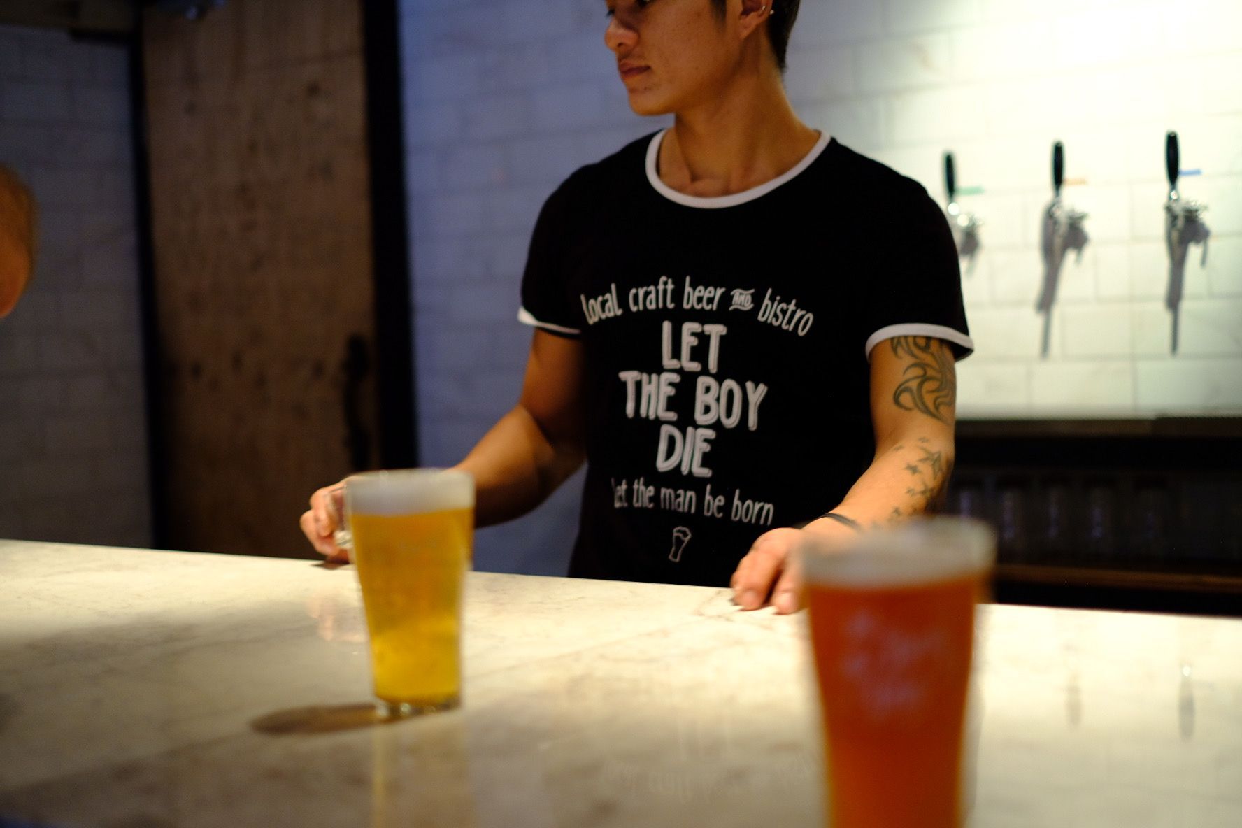 Bartender behind a bar with two beers in focus; wears a black shirt with text.