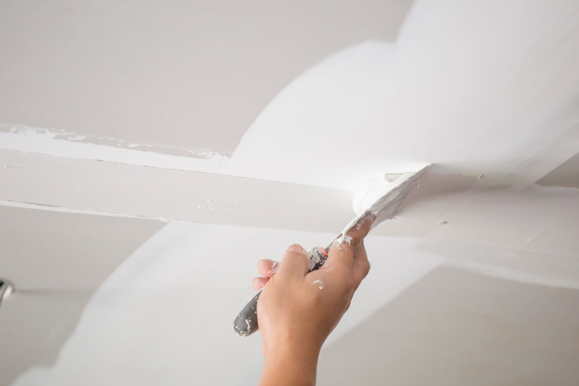 Hand using a putty knife to smooth white ceiling joint compound at a seam