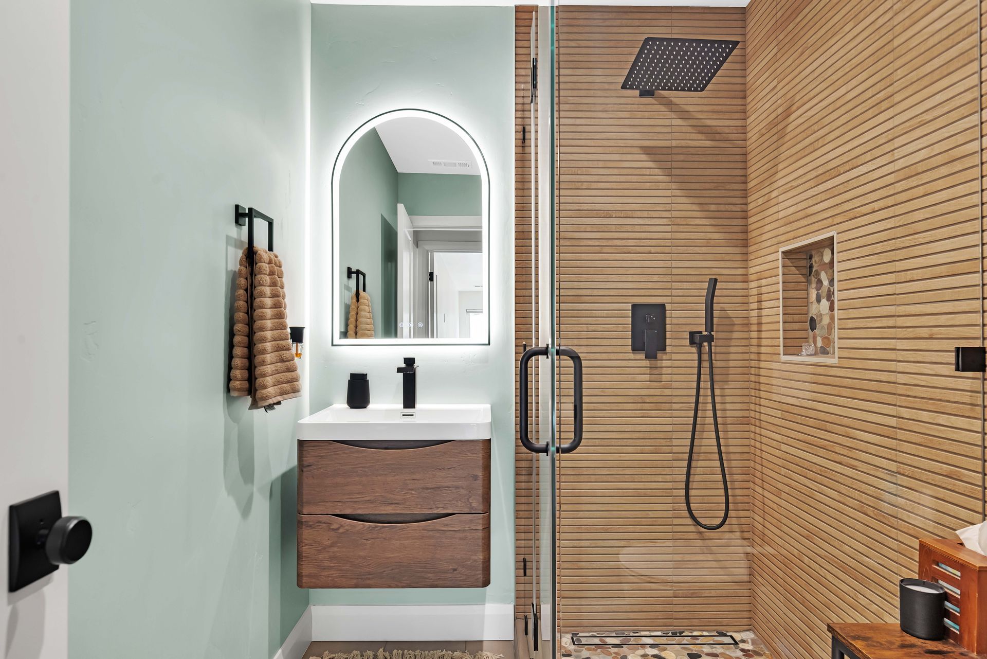 Bathroom with mint-green walls, wood-grain shower, floating vanity, arched mirror, black fixtures.