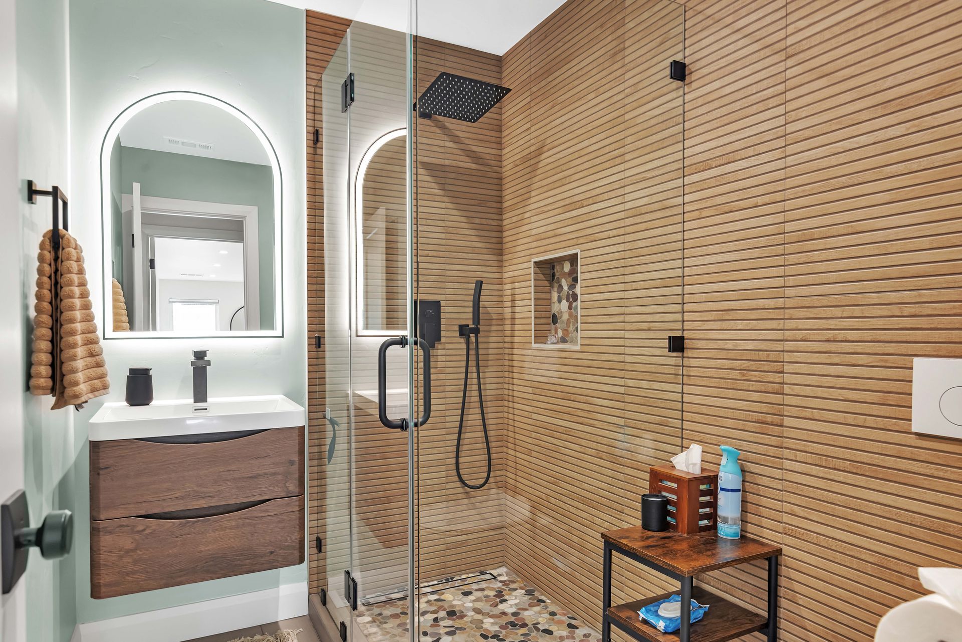 Modern bathroom with wood-look tile, glass shower, floating vanity, and arched mirror with halo lighting.