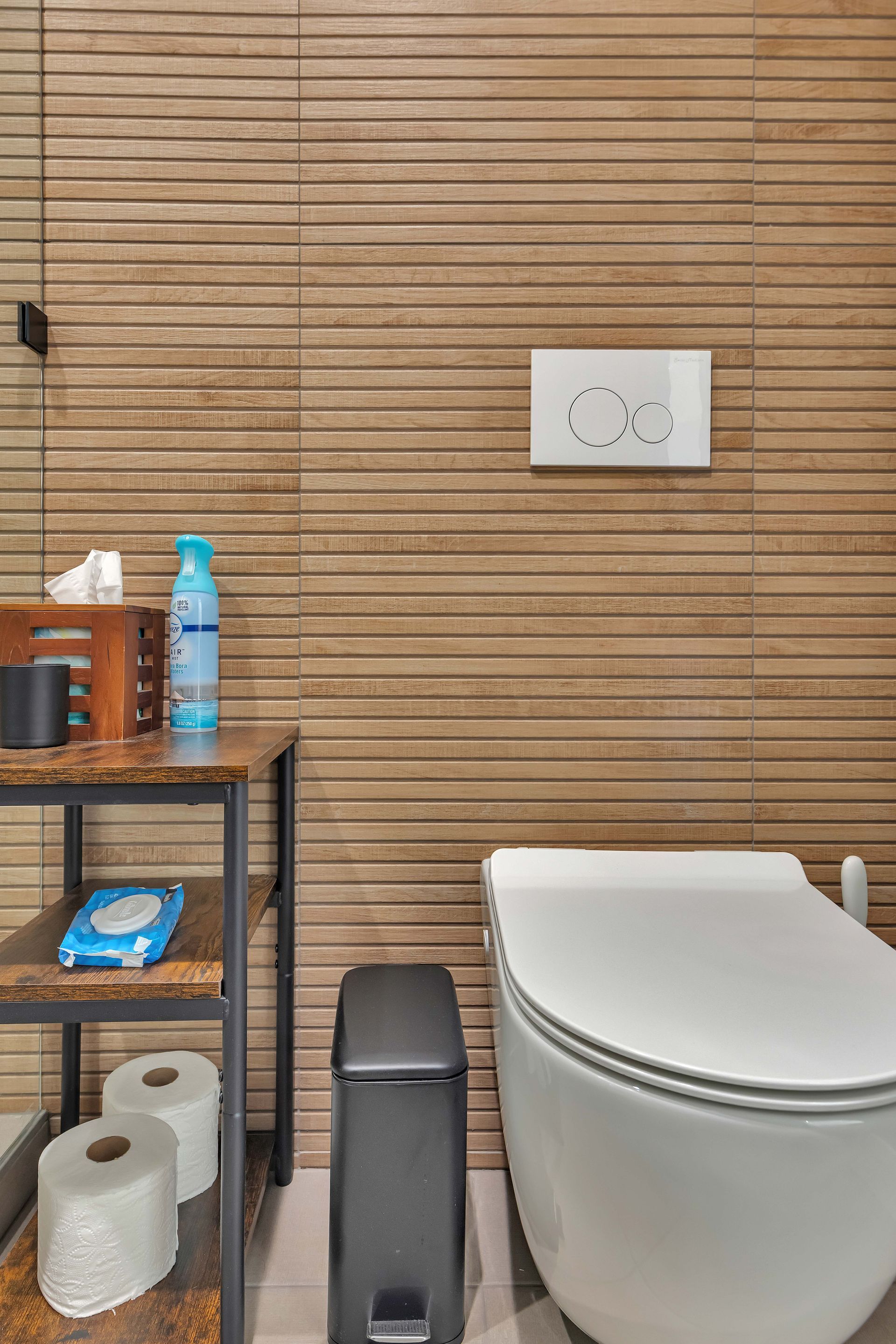 Bathroom with wood-look tile, toilet, shelf with supplies like wipes, tissue, spray, and toilet paper, and a black trash can.
