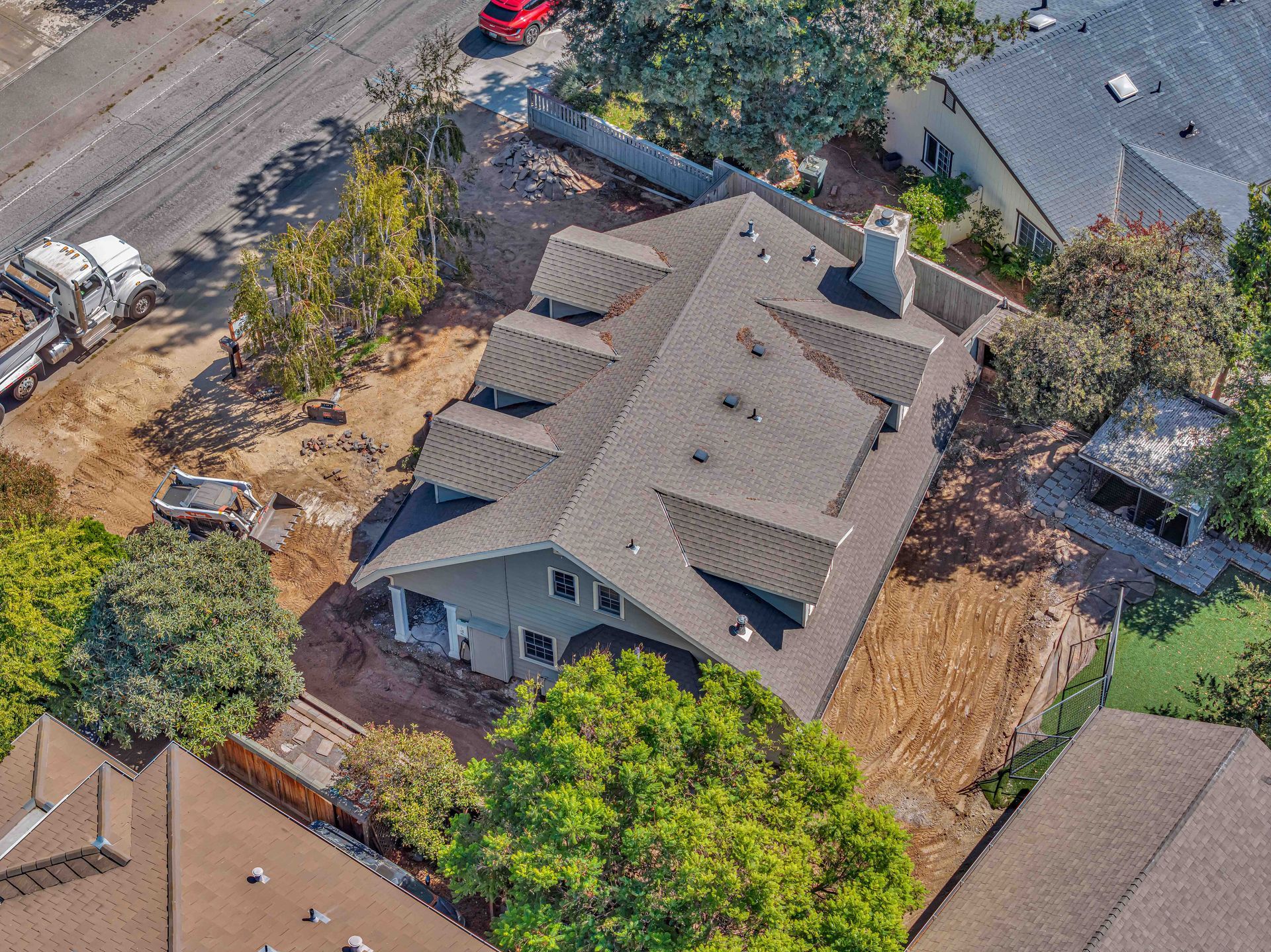 Aerial view of a gray house with a multi-level roof under construction, surrounded by brown dirt and some trees.