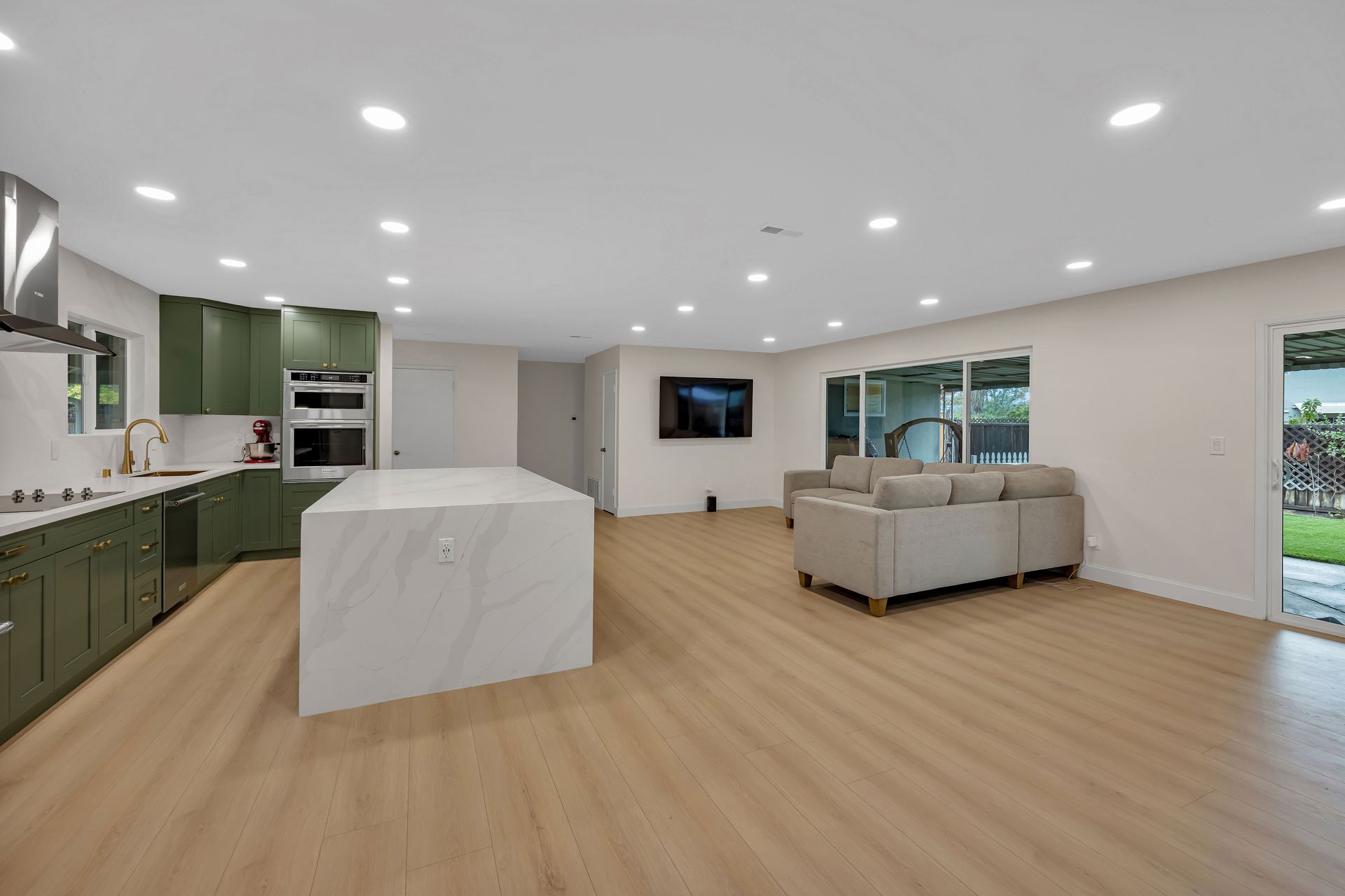 Open-concept kitchen and living area with olive green cabinets, white island, and light wood flooring.