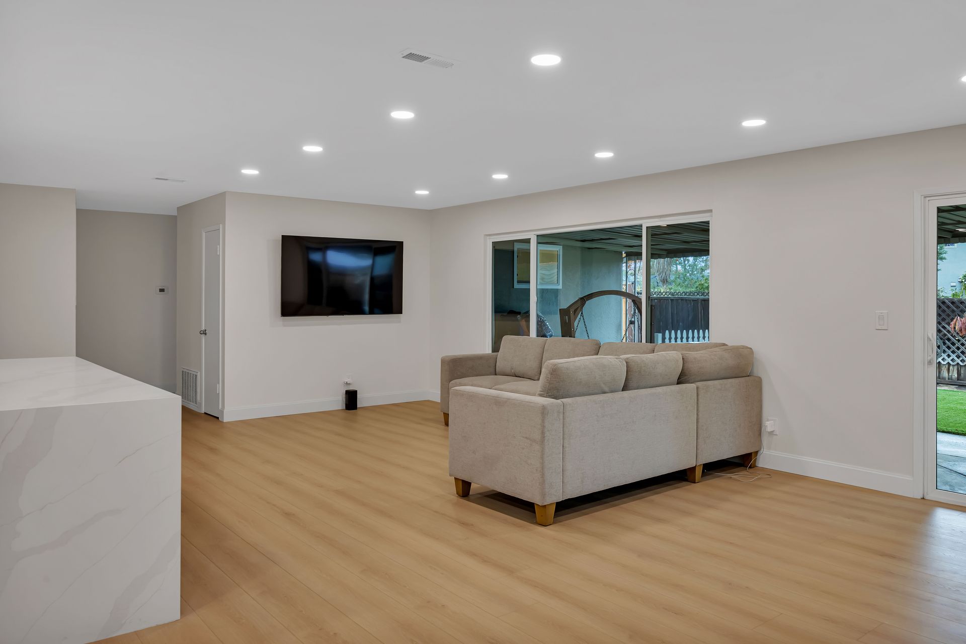 Empty living room with light wooden floors, white walls, and a mounted TV. Sliding glass door to a backyard.