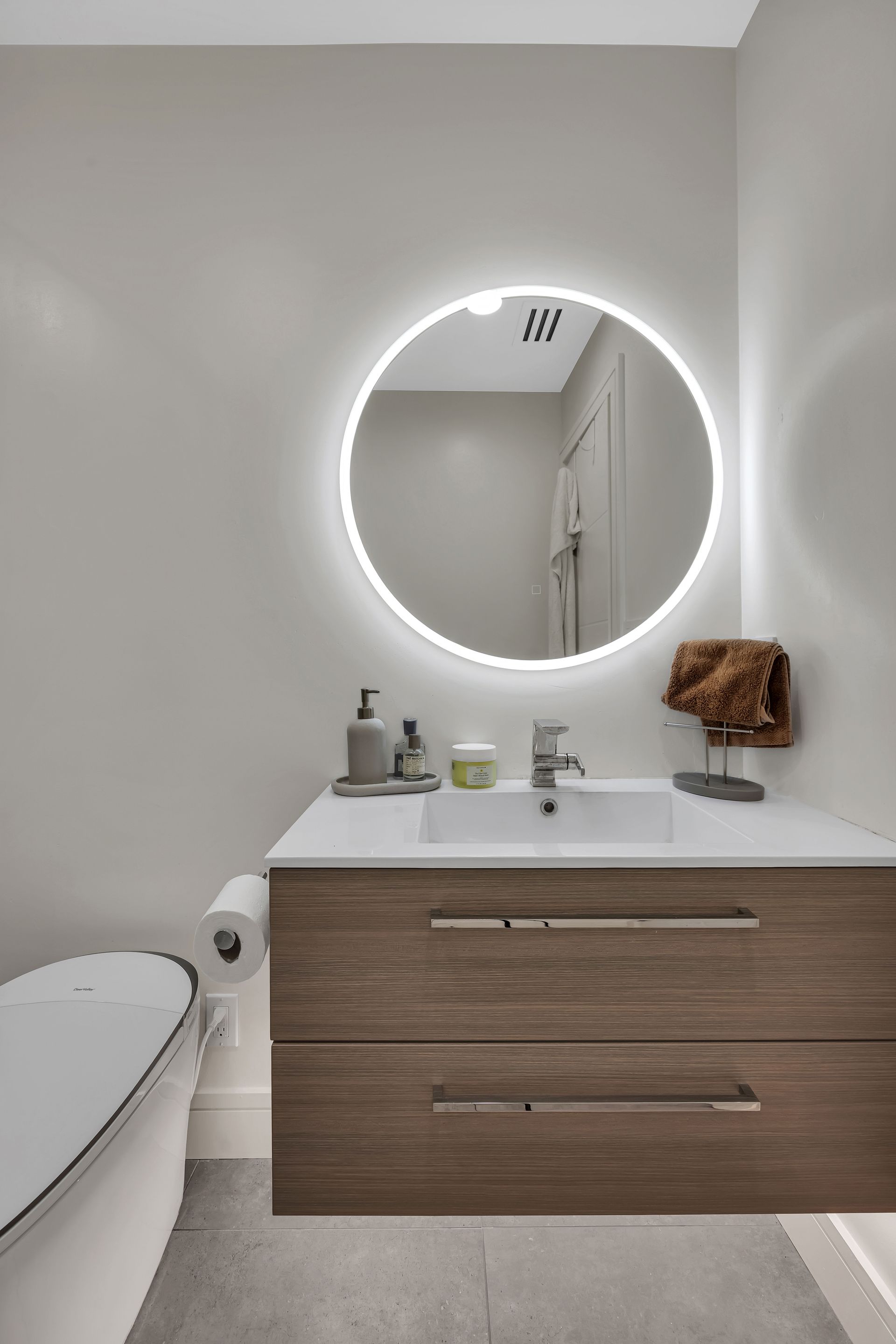 Modern bathroom with wood vanity, round mirror with light, and white toilet.