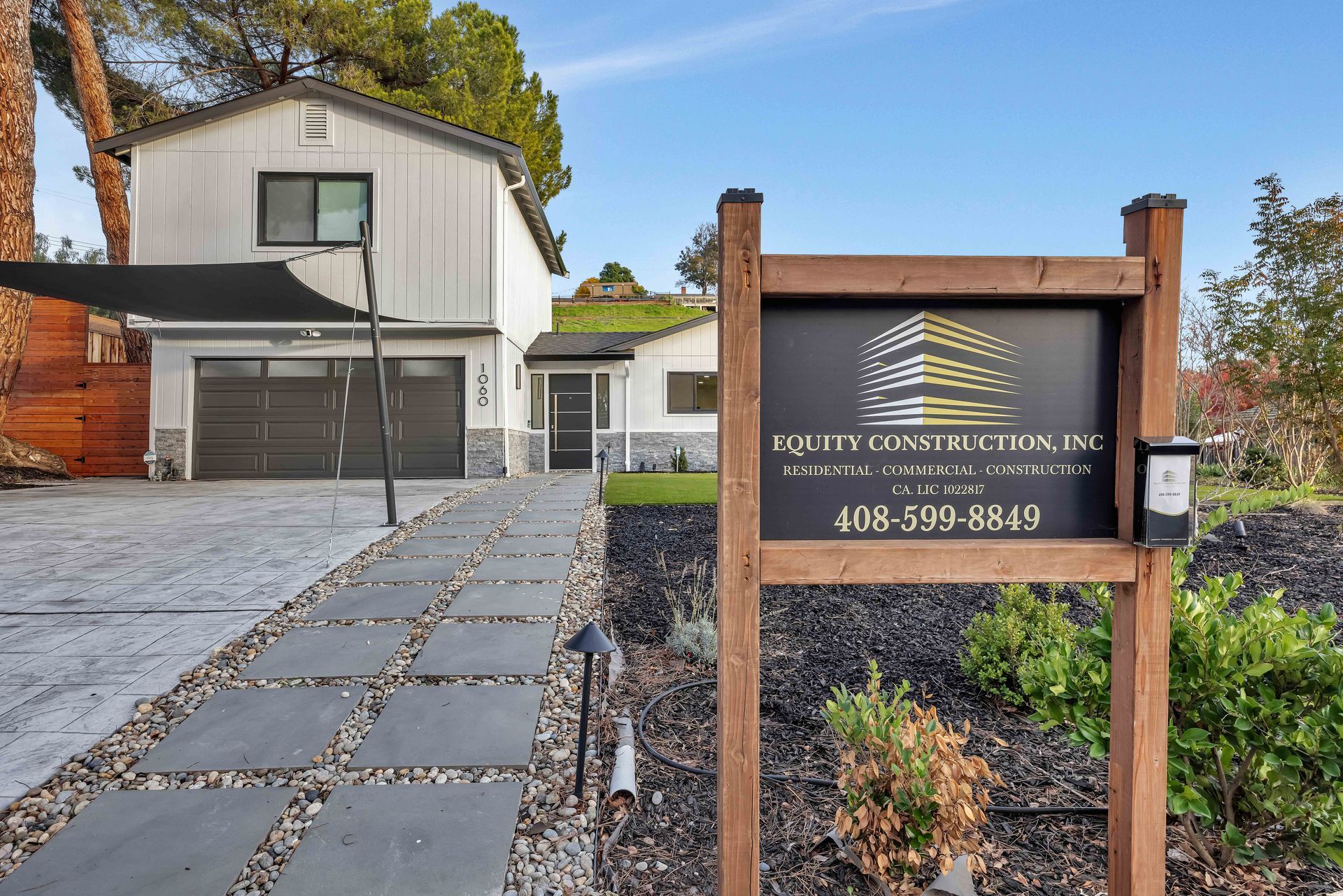 House exterior with sign. Gray and white house with a dark driveway and a brown wooden sign with a phone number.