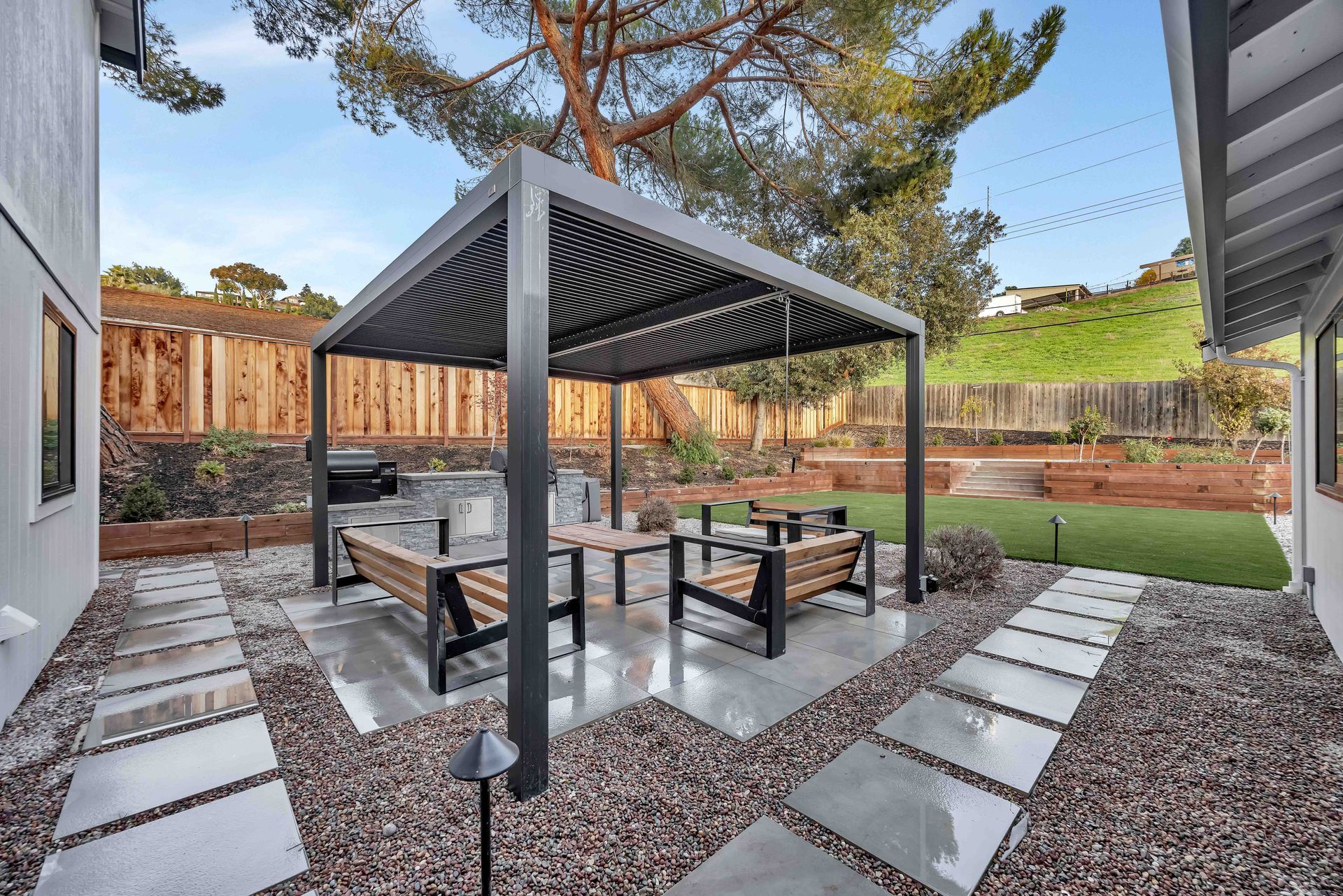 Outdoor patio with a pergola, seating, and a grill, surrounded by a fence and landscaping.