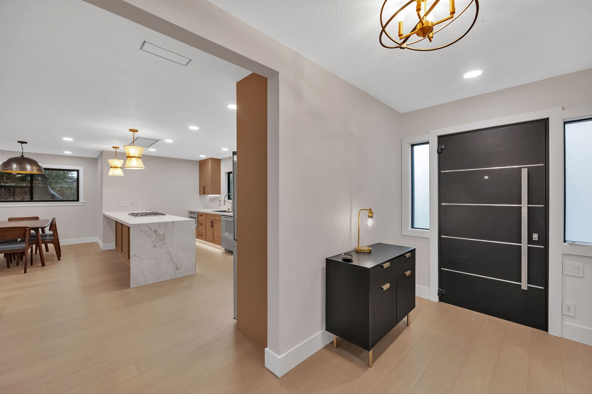 Modern home entryway with black door, console table, and light wood flooring. Kitchen visible in the background.